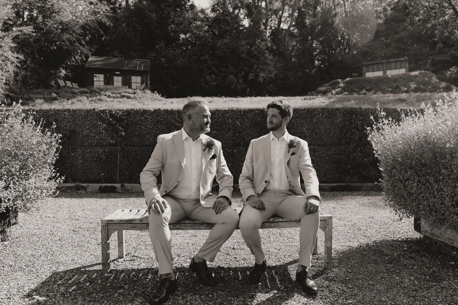 Black and white photo of two grooms in light suits sitting together on a garden bench, sharing a joyful moment.