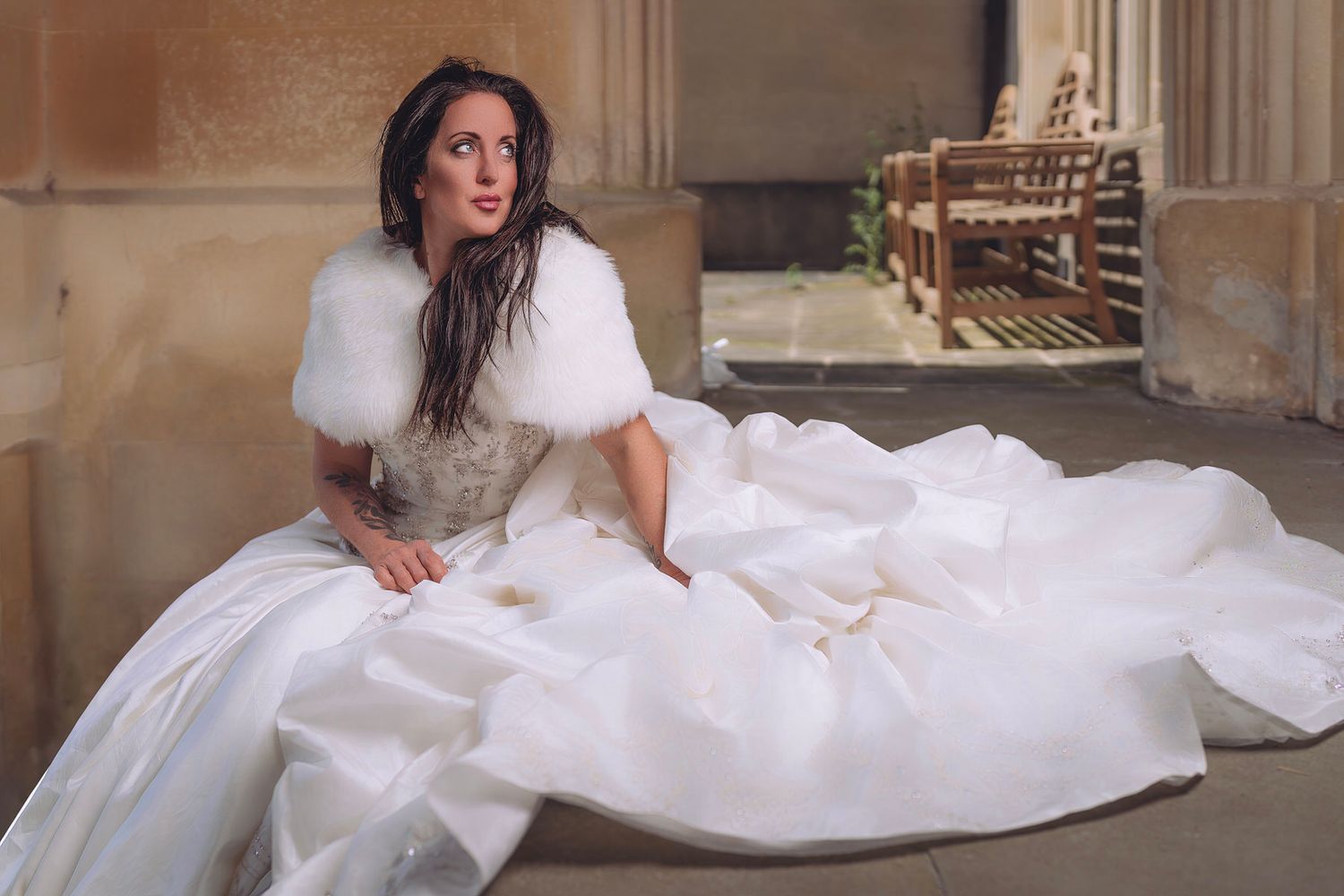 Bride in elegant white gown with fur shawl poses gracefully on stone floor in grand architectural setting.
