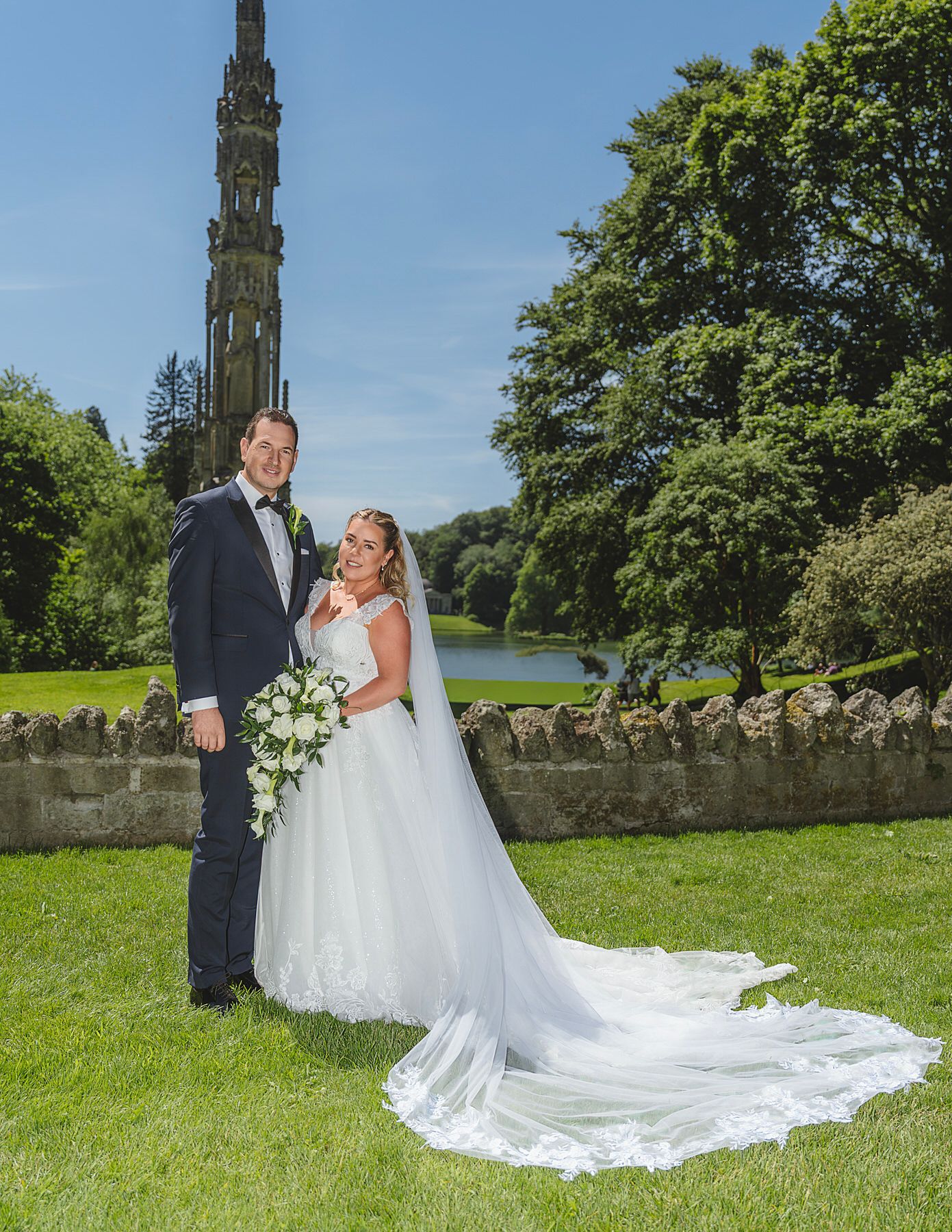 Bride and groom pose on sunny lawn with long cathedral veil and Gothic monument in background.