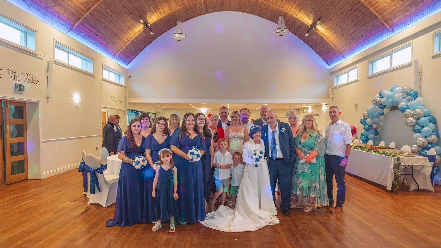 Wedding party posing together in a decorated hall with blue balloons, bridesmaids in navy dresses flanking the bride and groom.