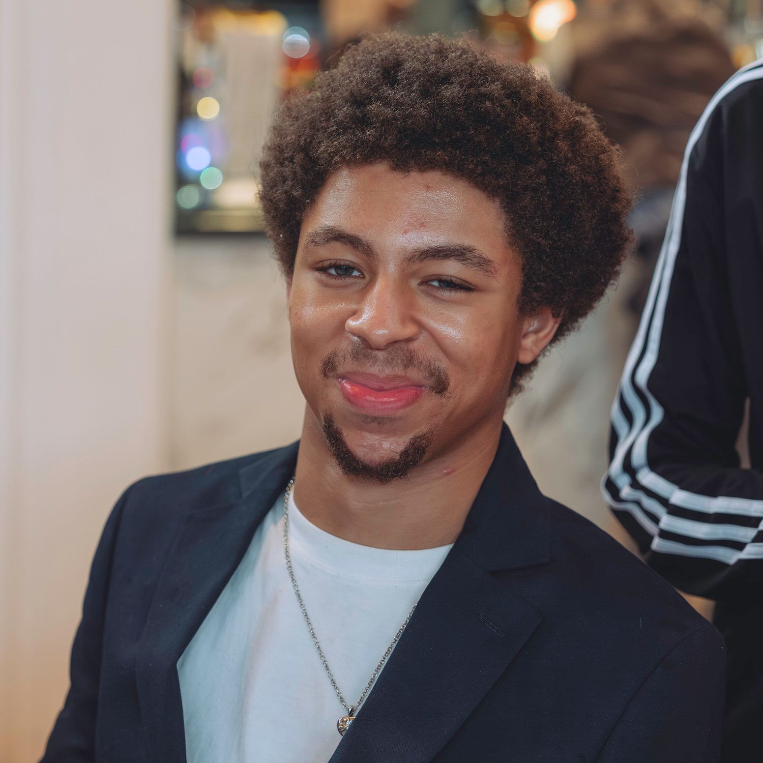 Young man with curly hair smiling warmly, wearing a black blazer and white t-shirt with a silver necklace.