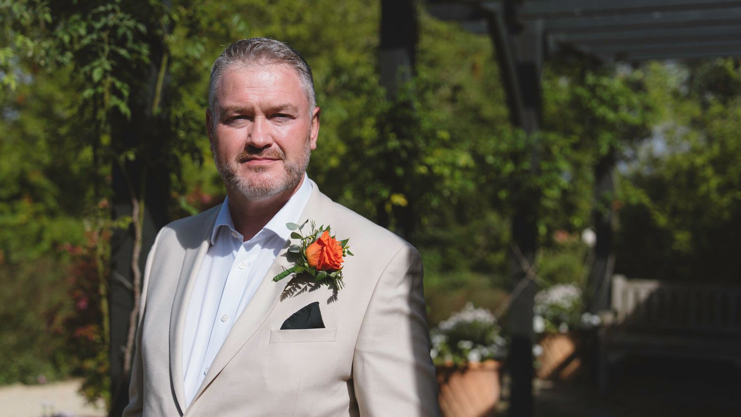Man in beige suit with orange boutonniere posing outdoors at a wedding ceremony.
