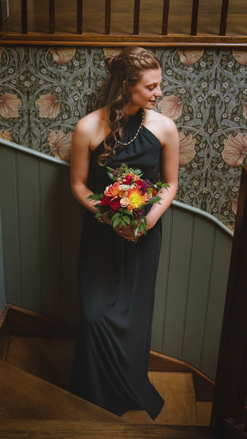 Bridesmaid in black halter gown holding vibrant bouquet on staircase with floral wallpaper backdrop.