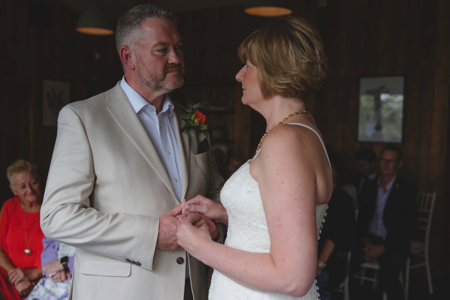 Bride and groom exchanging rings during indoor wedding ceremony, groom in beige suit, bride in white lace dress.