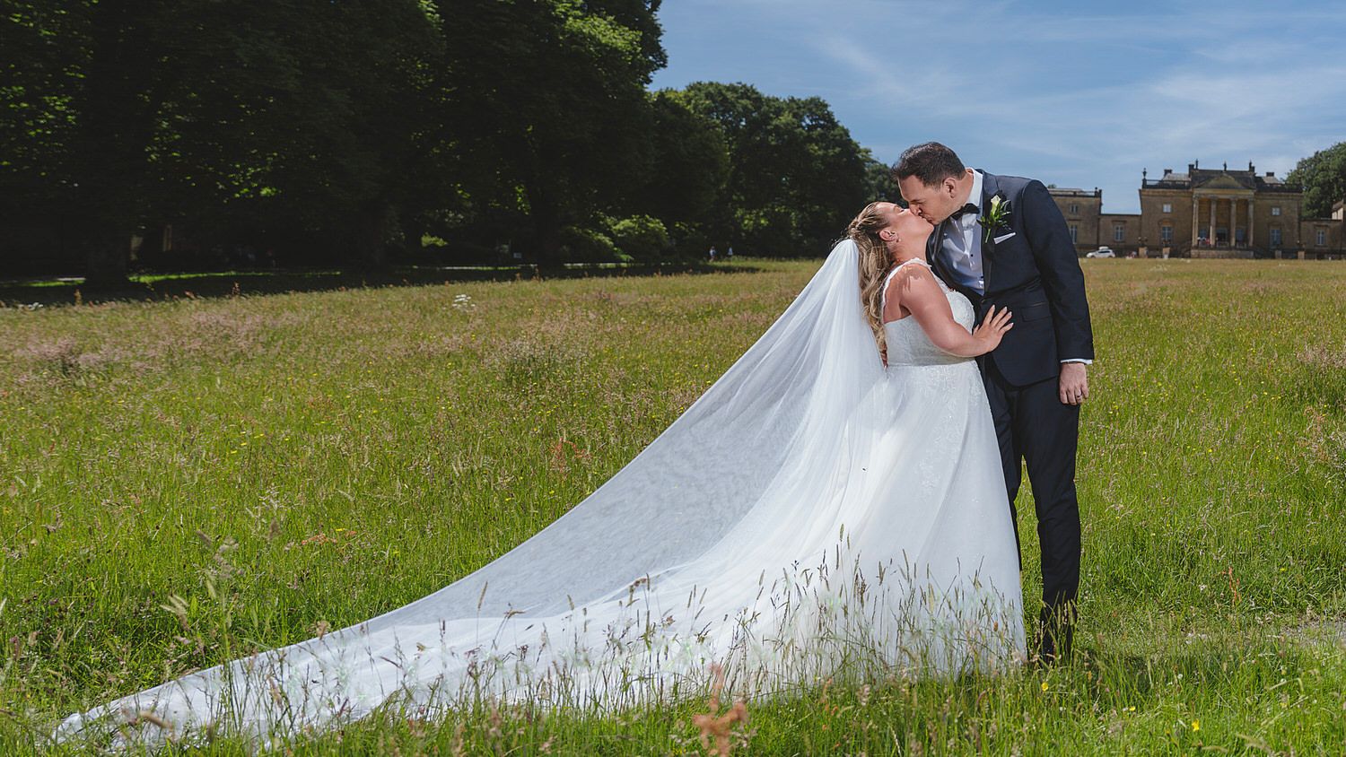 Bride and groom share a romantic kiss in a lush green meadow, her long cathedral veil trailing behind them.