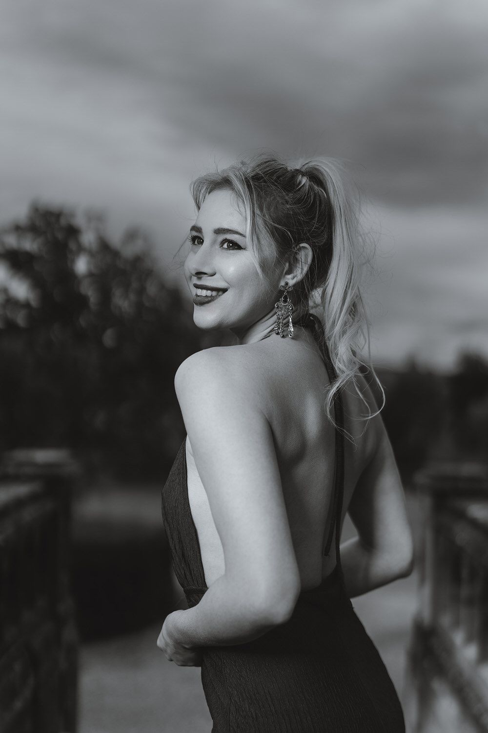 Black and white portrait of a smiling blonde woman in a backless dress, posing outdoors at dusk.
