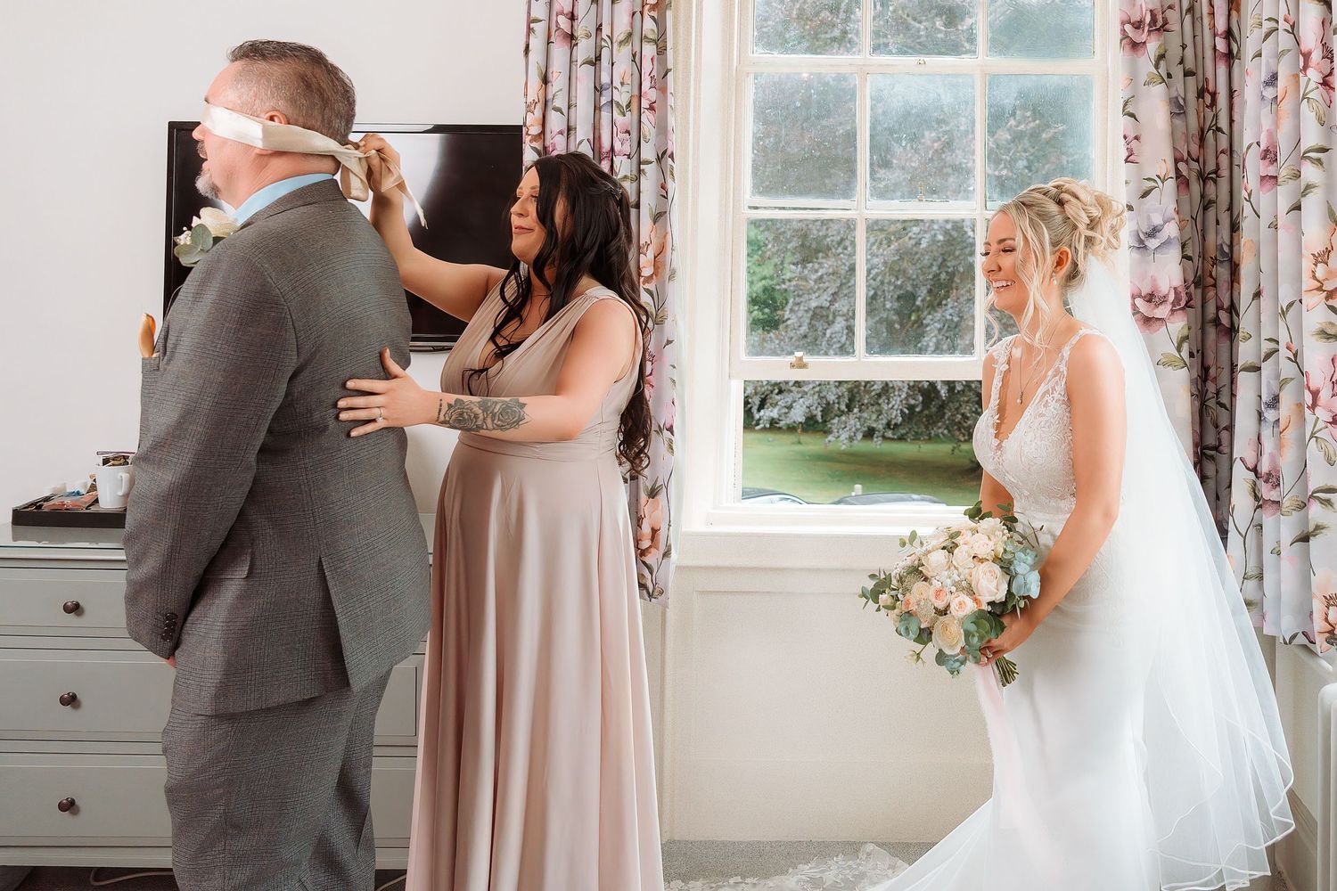 Bride in white gown holds bouquet while bridesmaid adjusts groomsman's tie near a bright window.