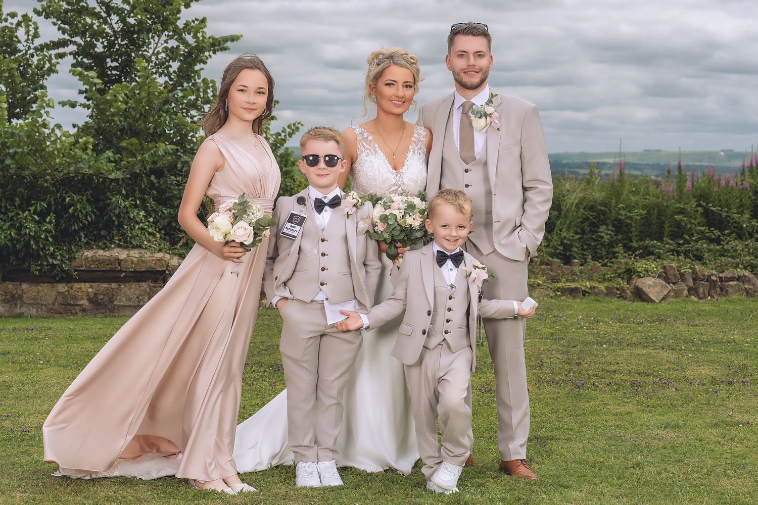 Wedding party posing outdoors, bride in white gown with bridesmaids in blush pink and groomsmen in tan suits.