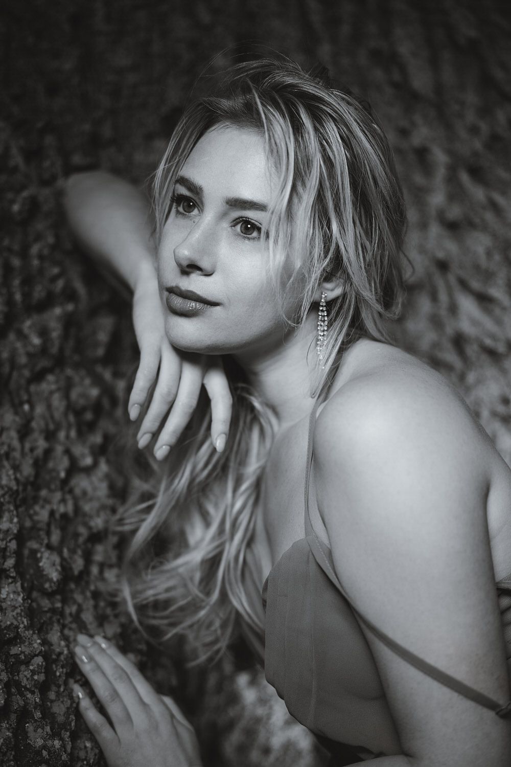 Black and white portrait of a blonde woman leaning against a tree, wearing a spaghetti strap top and chandelier earring.