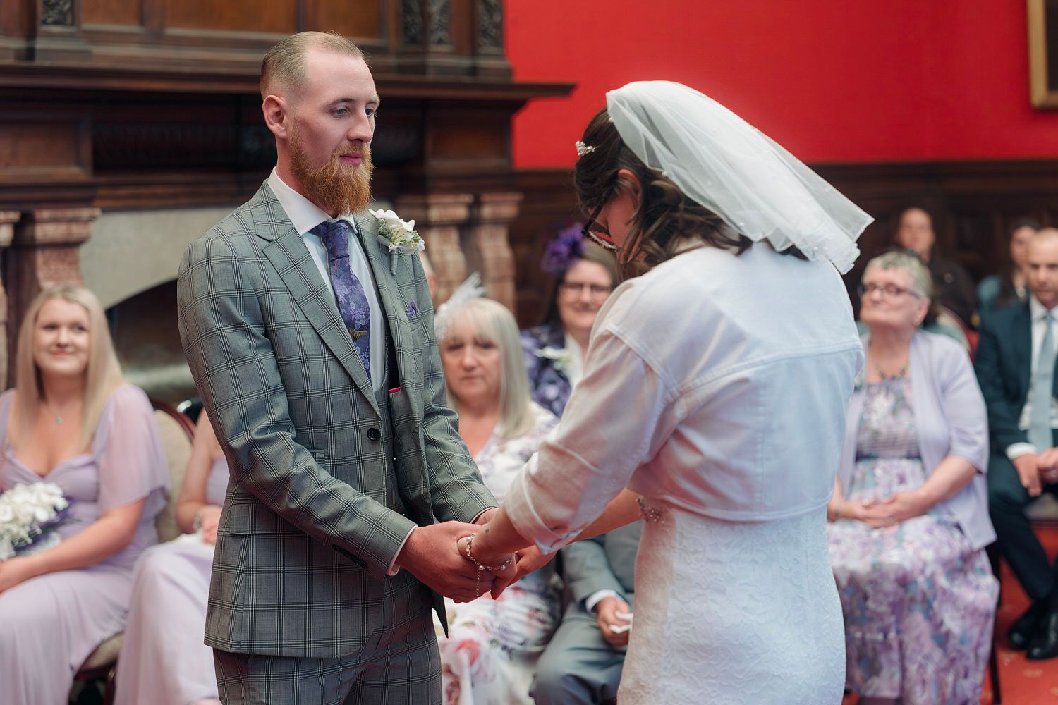 Groom with red beard in grey suit holds hands with bride in white veil during wedding ceremony.