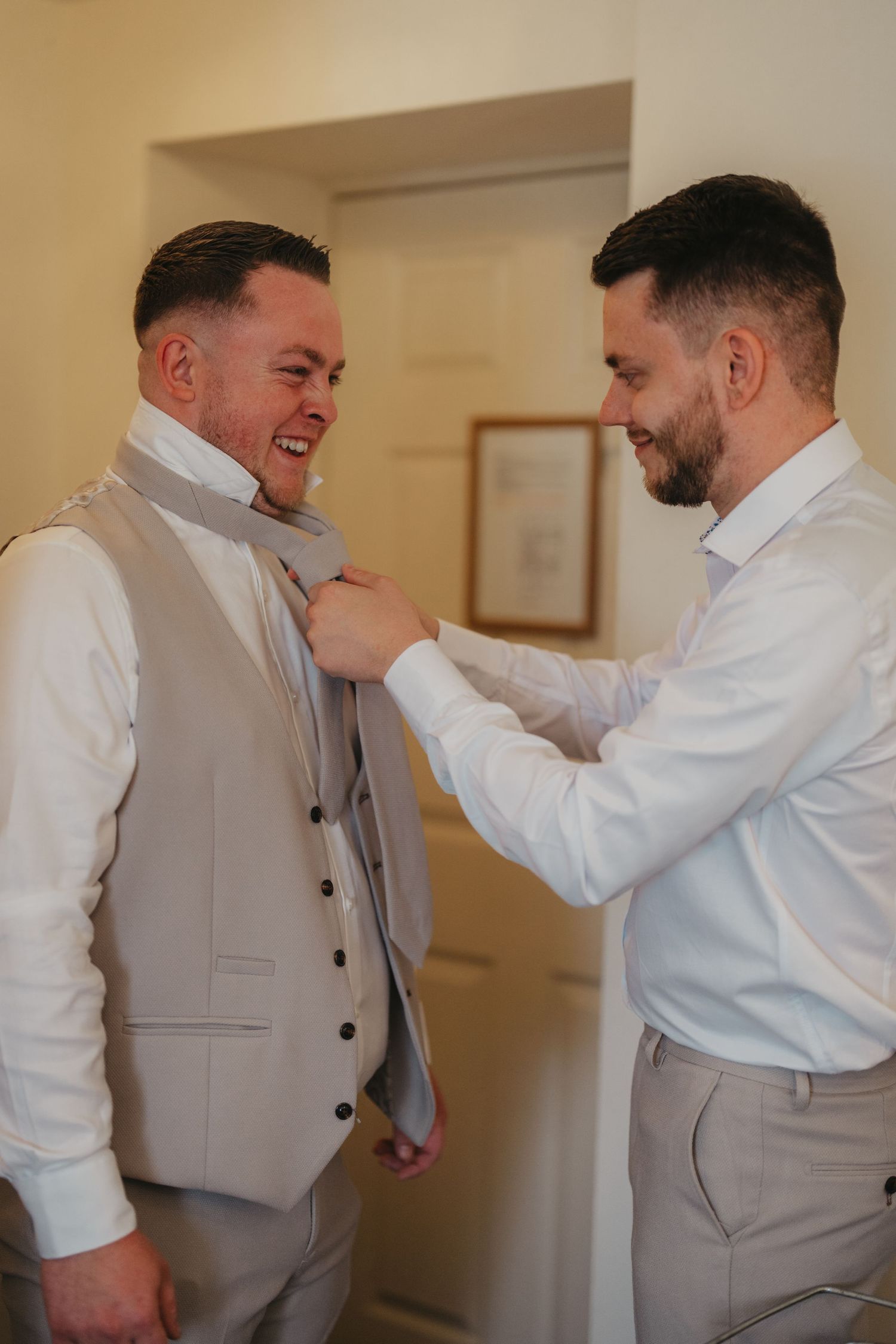 Groomsman helping groom adjust grey tie while both smile during wedding preparations indoors.