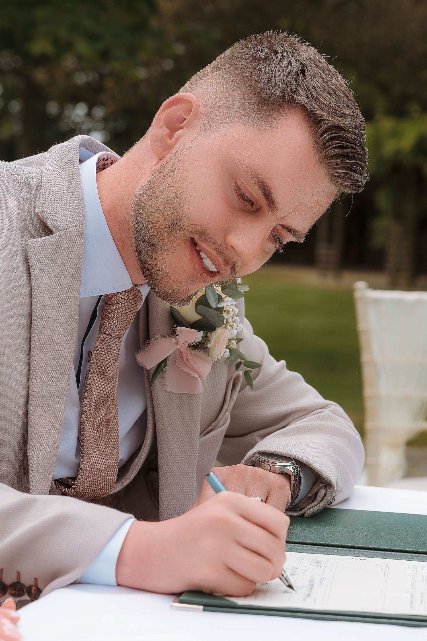 Groom in beige suit signing wedding documents outdoors, wearing boutonniere and tie.