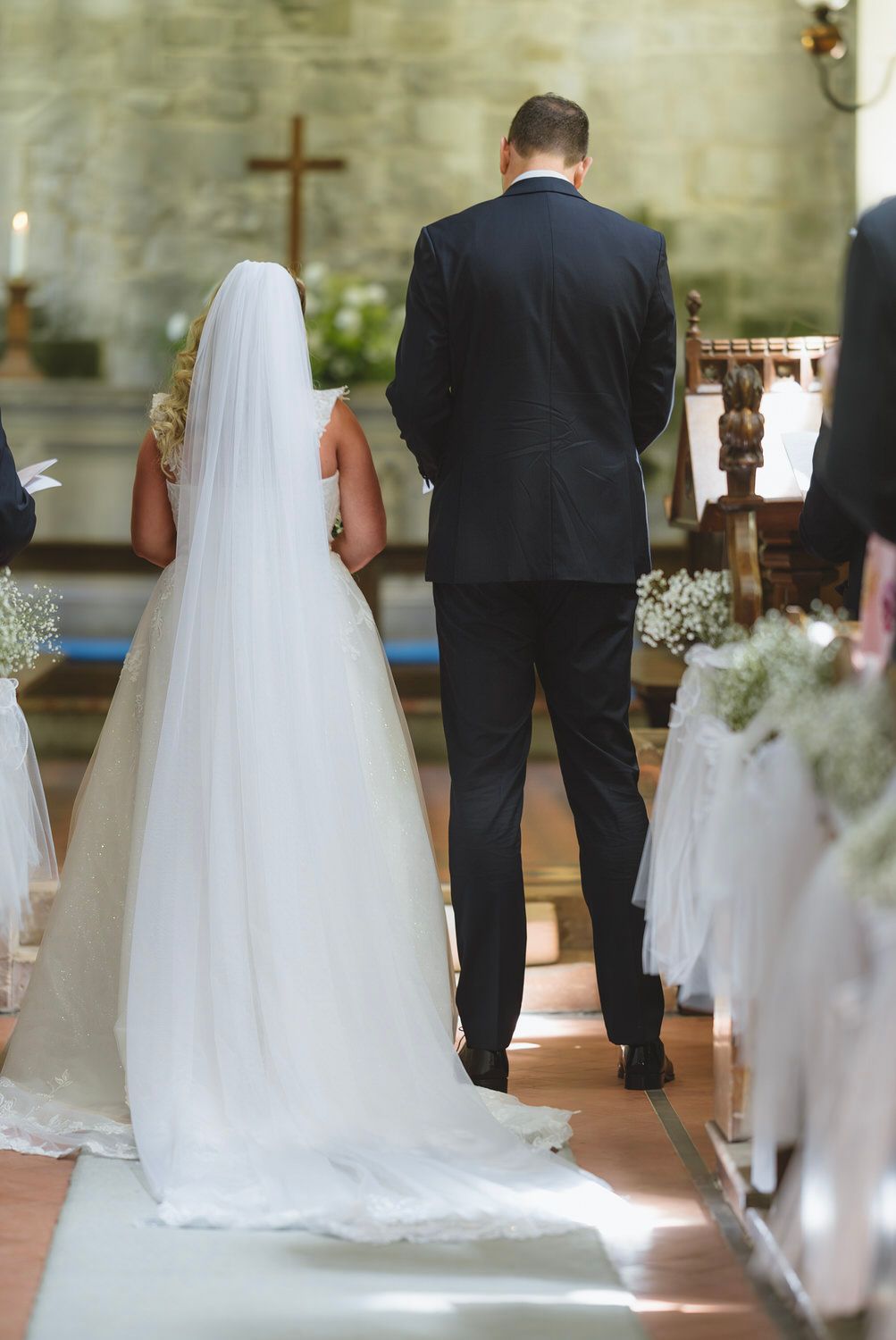 Bride and groom standing at church altar during wedding ceremony, viewed from behind, with cross and floral decorations.