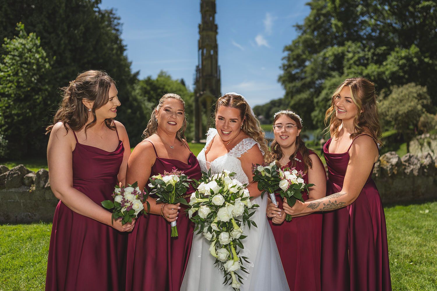 Bride in white gown with four bridesmaids in burgundy dresses holding white bouquets outdoors on a sunny day.