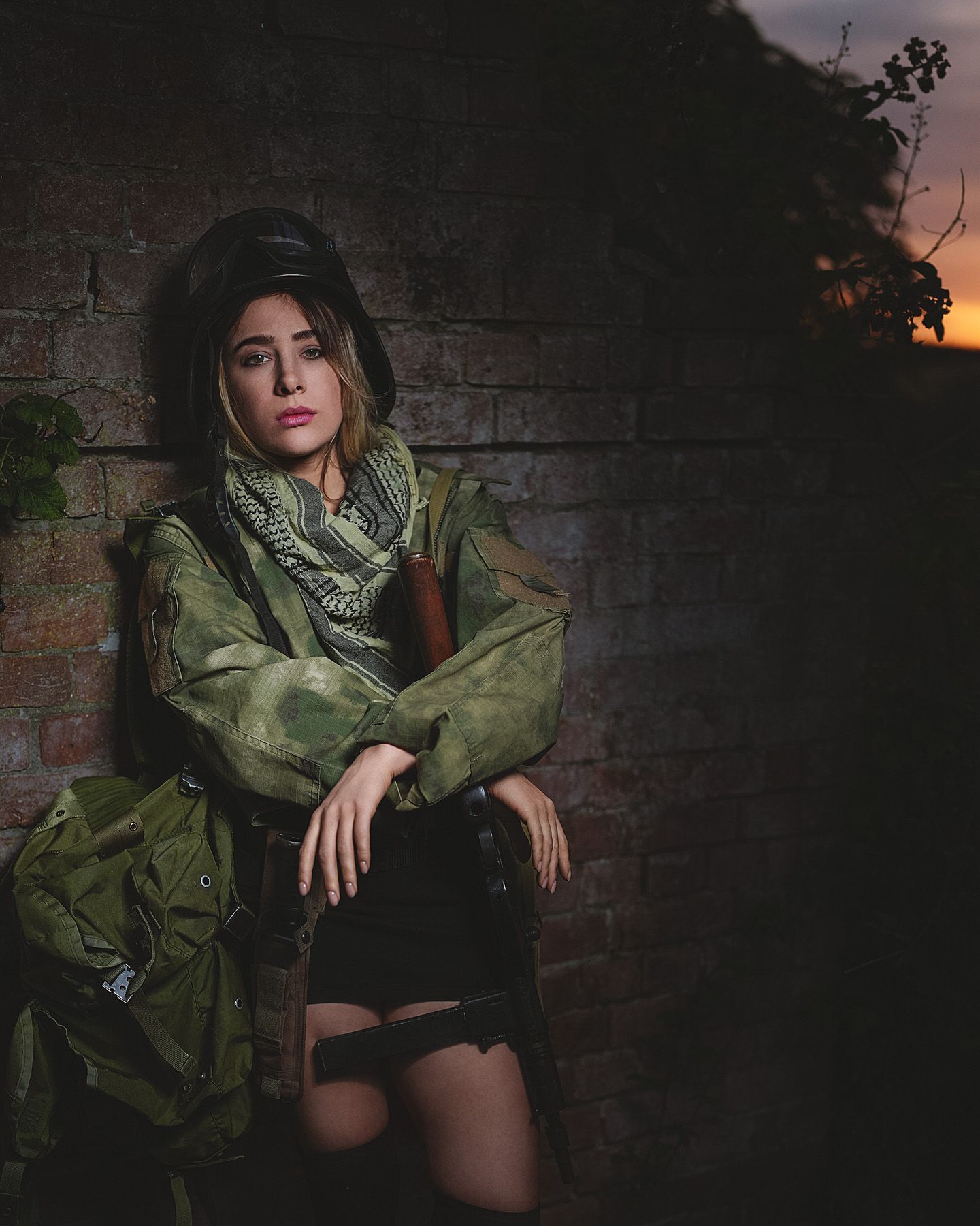 Woman in military camouflage jacket leaning against brick wall at sunset with backpack.