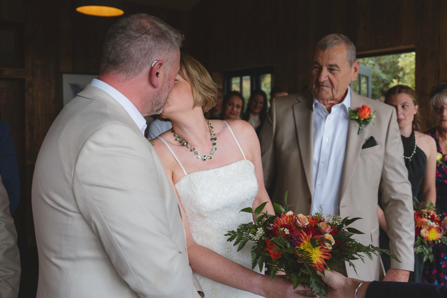 Bride and groom share a first kiss at rustic wedding ceremony while older gentleman watches nearby.