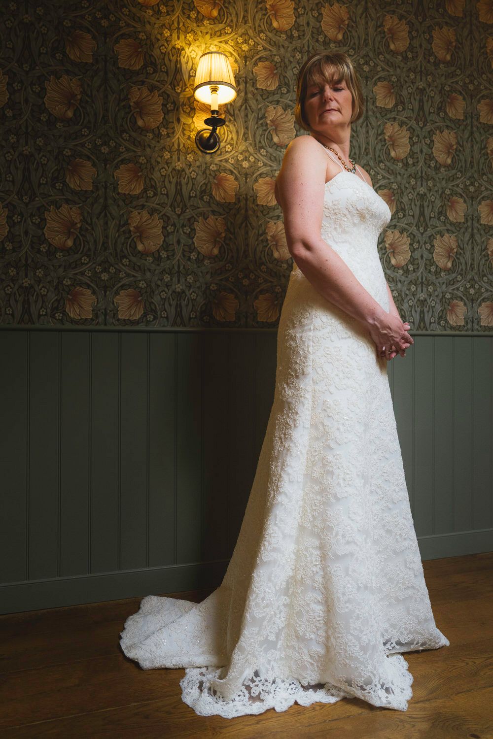 Bride in white lace wedding gown with train poses in room with ornate wallpaper and wall sconce lighting.