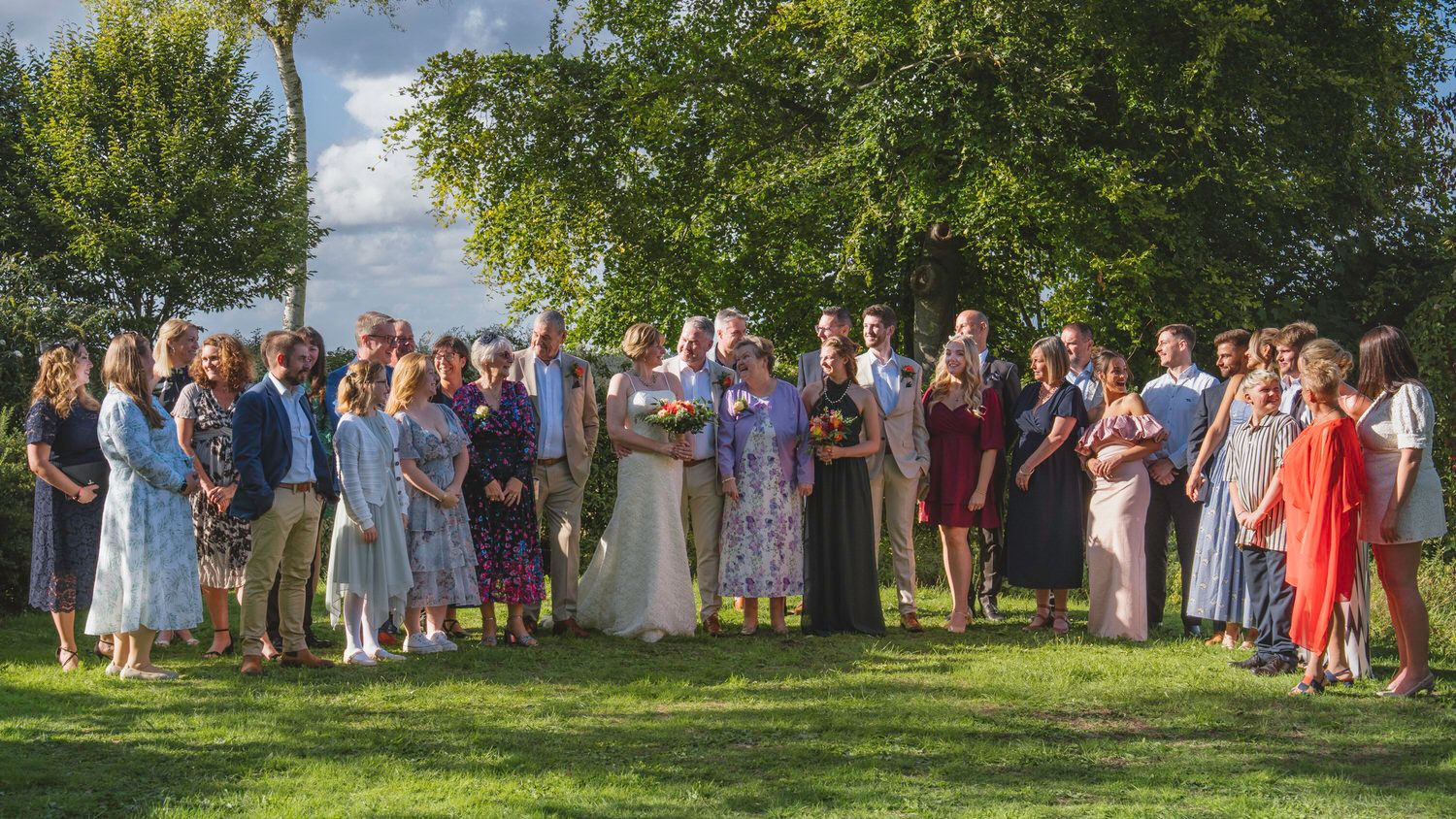 Large wedding party group photo outdoors on green lawn with trees, bride in white gown surrounded by guests.