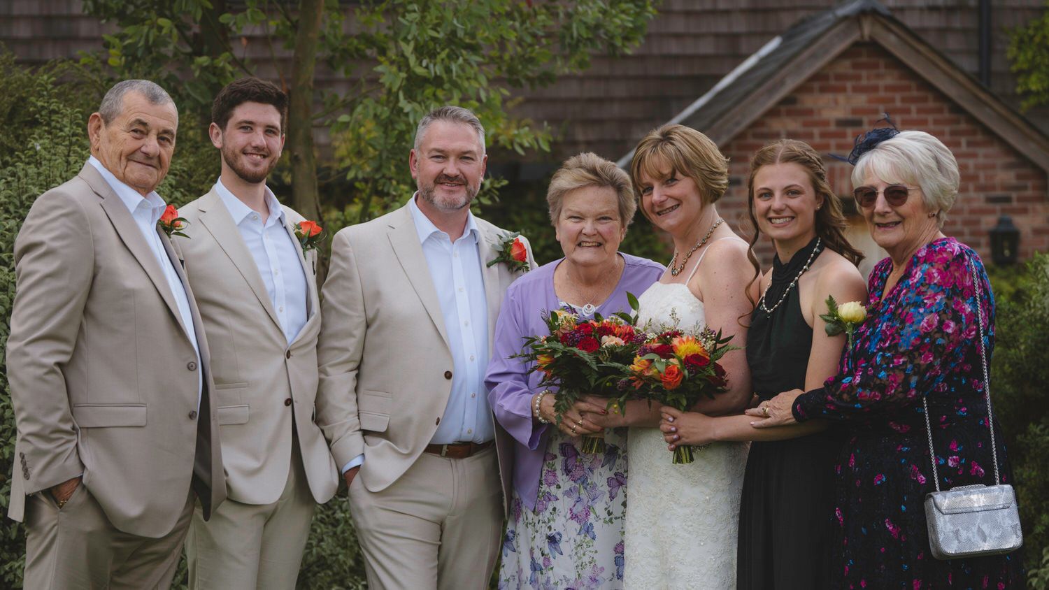 Wedding group photo with bride holding colorful bouquet, posing with family outdoors near brick building.