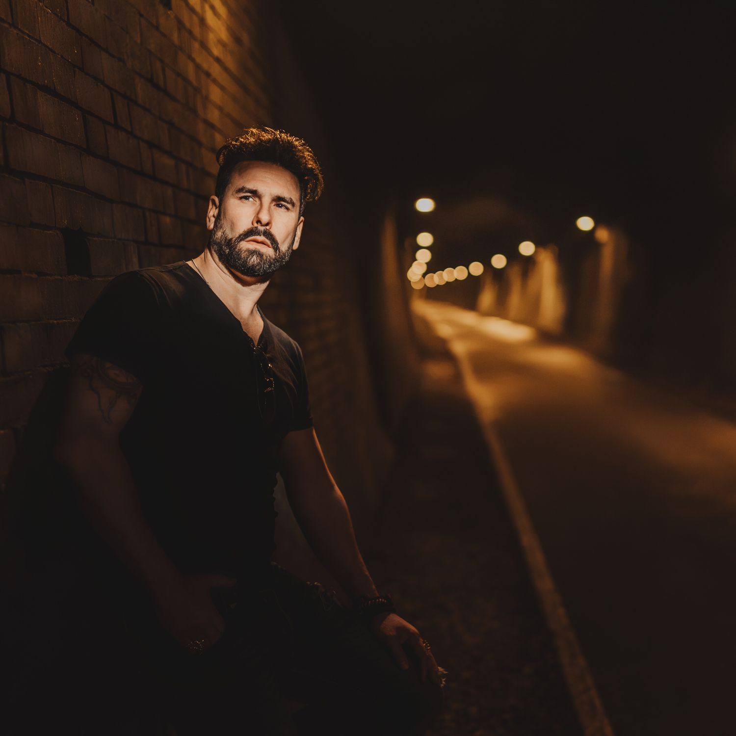 Bearded man in black t-shirt leaning against brick wall in illuminated tunnel at night.