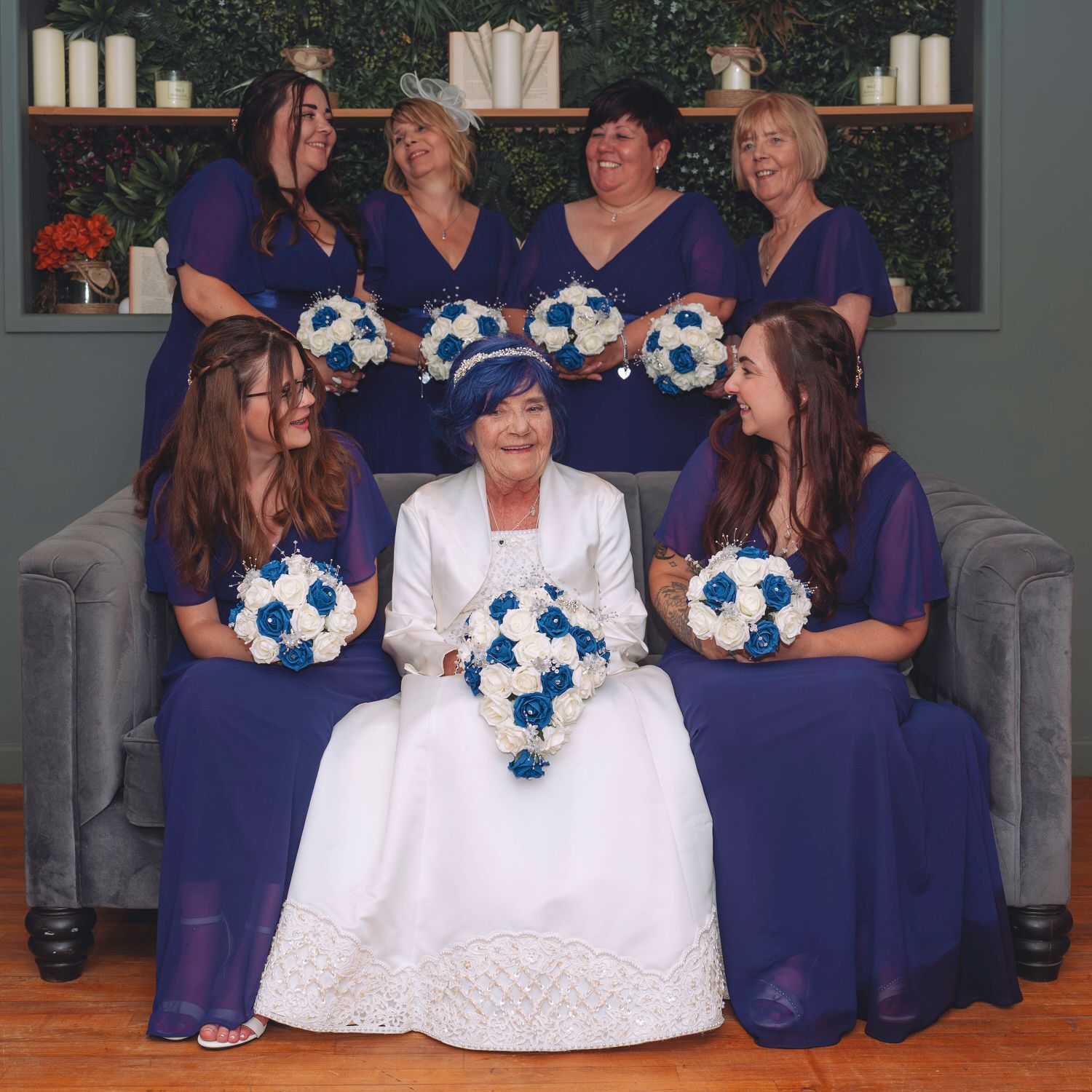 Bride in white dress sits surrounded by bridesmaids in royal blue gowns holding blue and white bouquets.