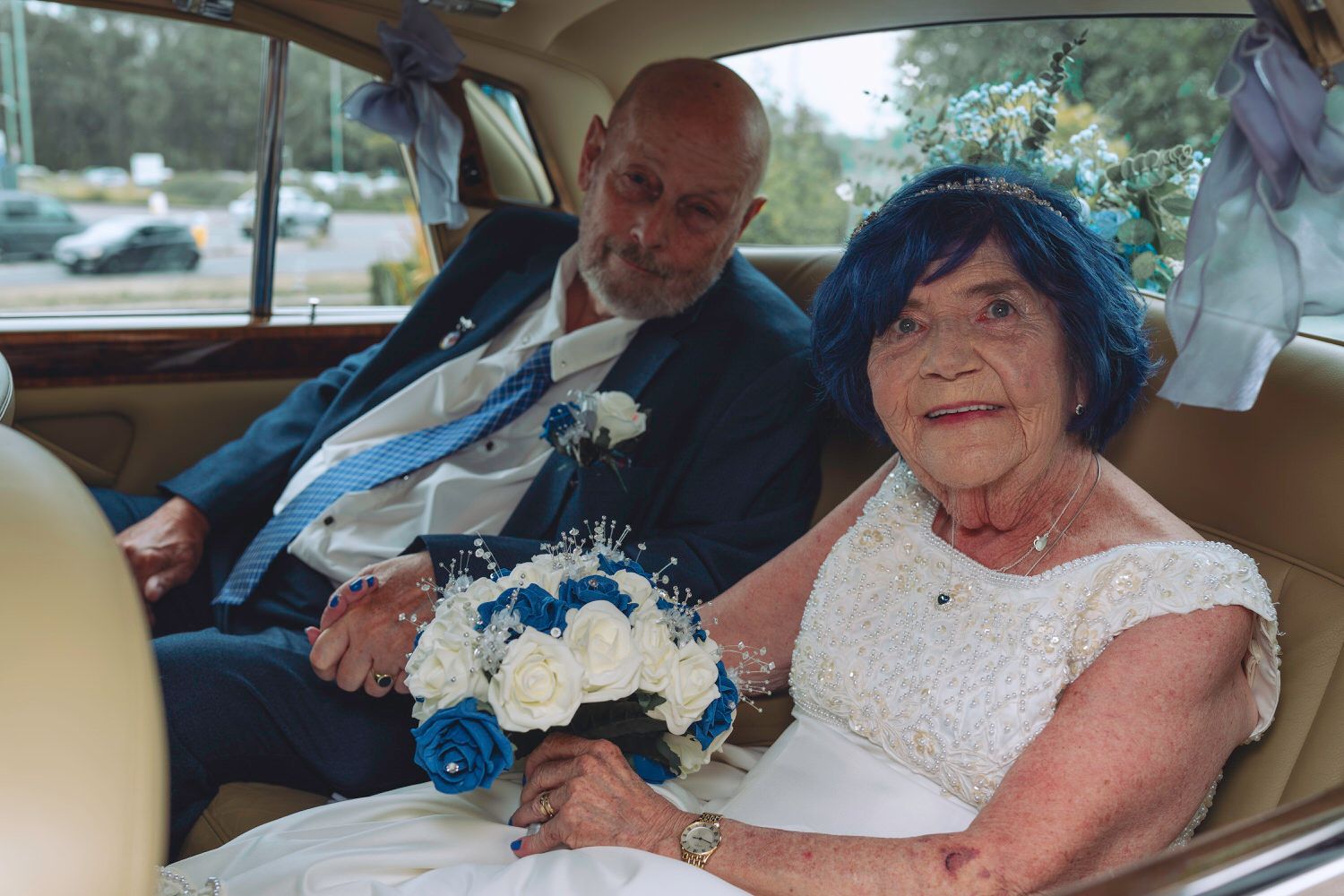 Elderly bride in blue hat holding blue and white bouquet sits with groom in classic car on wedding day.