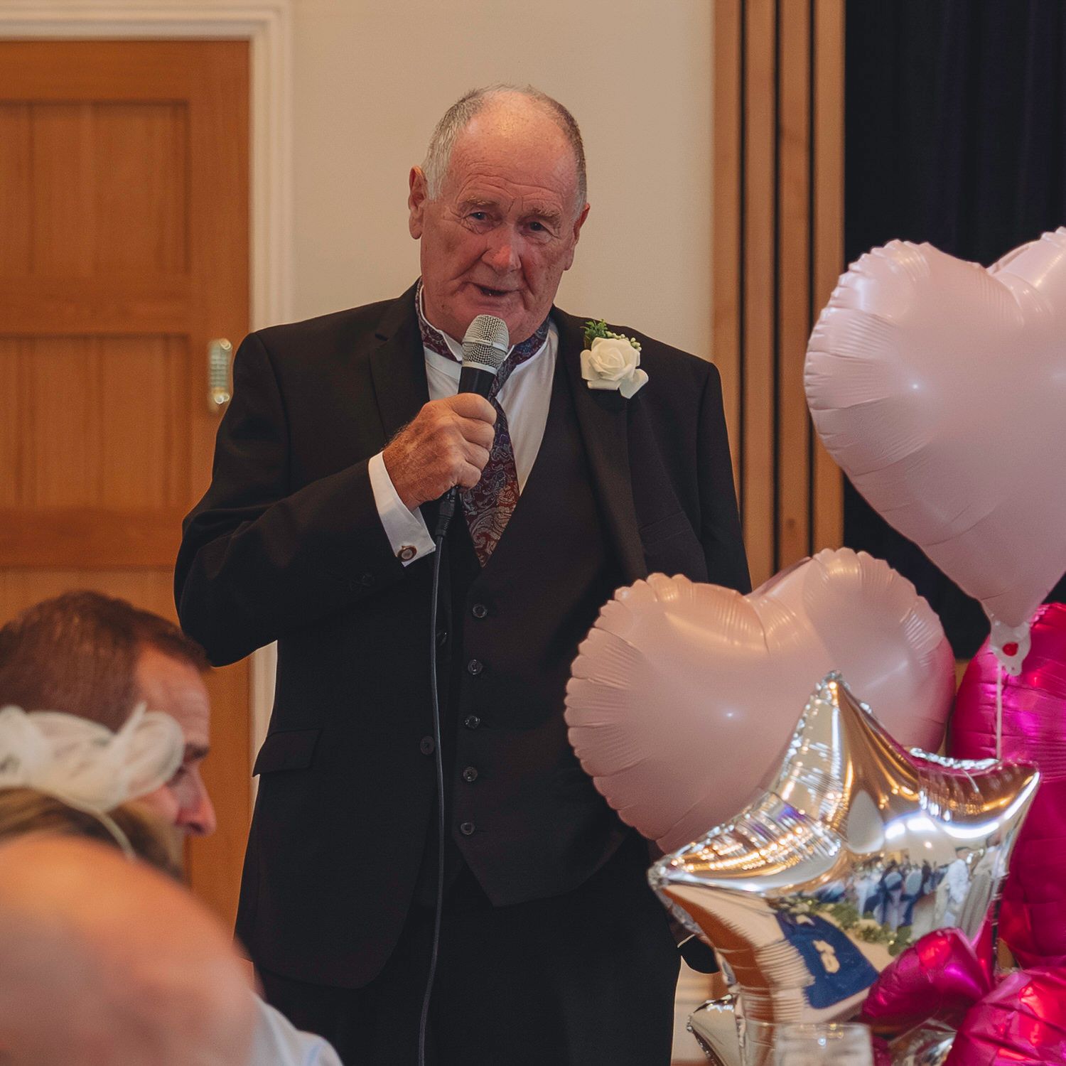 Elderly man in suit giving speech with microphone at wedding reception, pink heart balloons in foreground.