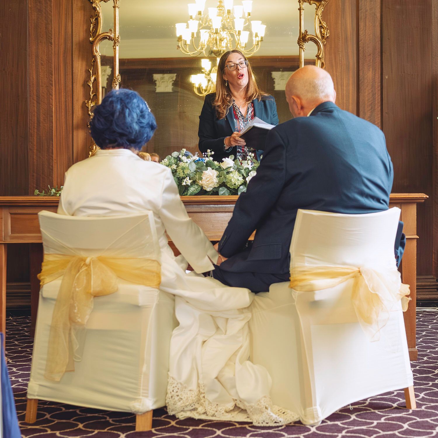 Couple holding hands during intimate wedding ceremony with officiant reading from book at flower-adorned altar.