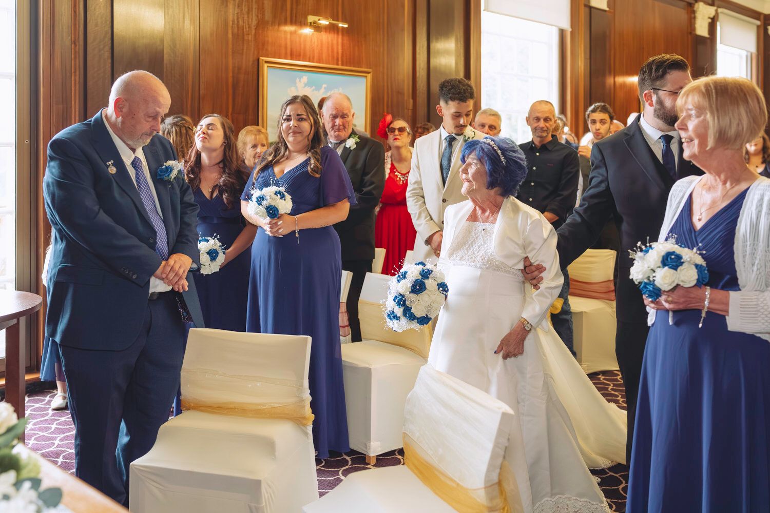 Wedding ceremony with bridesmaids in navy blue dresses holding bouquets as bride walks down the aisle.
