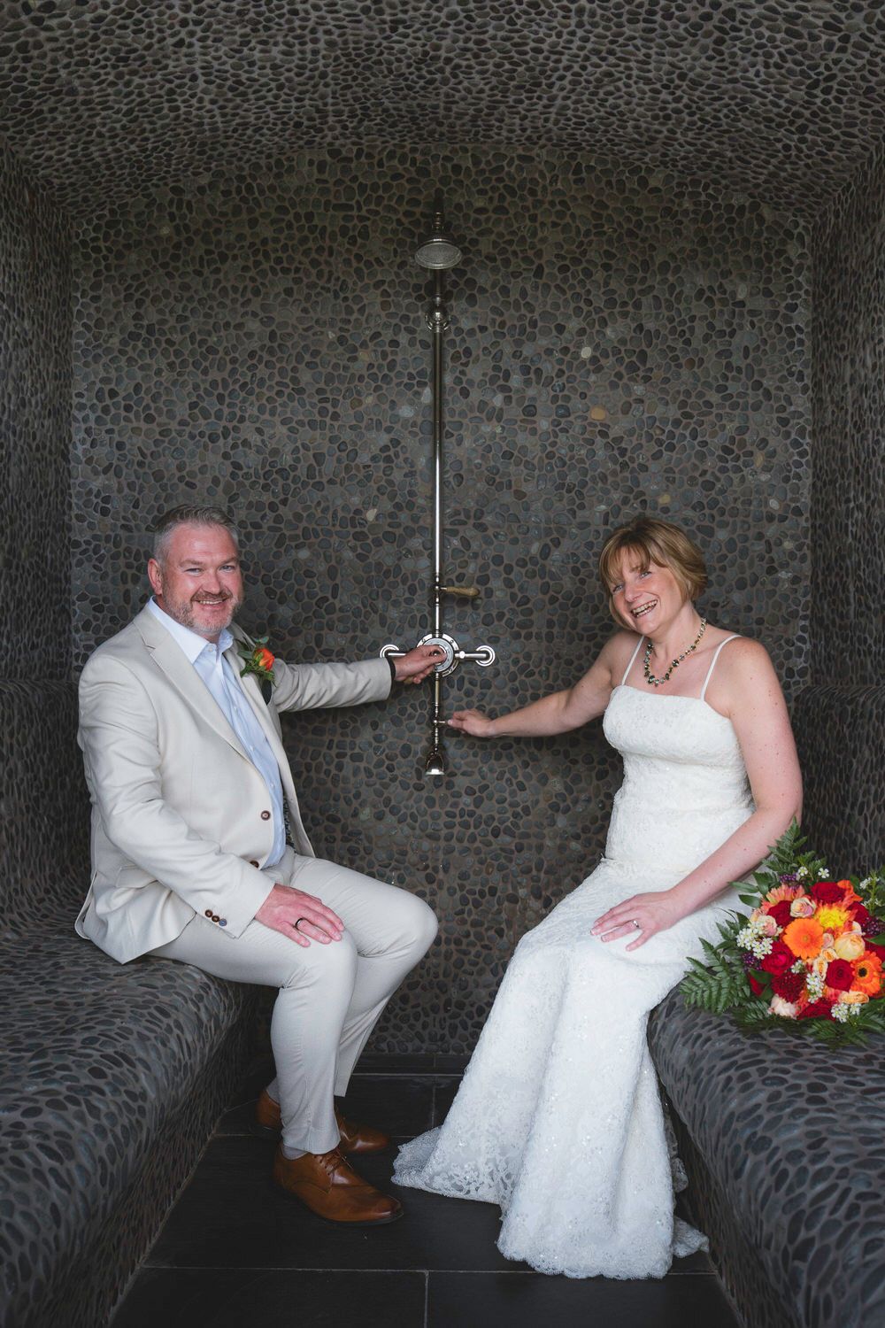 Bride and groom pose in a stone shower room, she holds a colorful bouquet, he wears a tan suit with floral boutonniere.