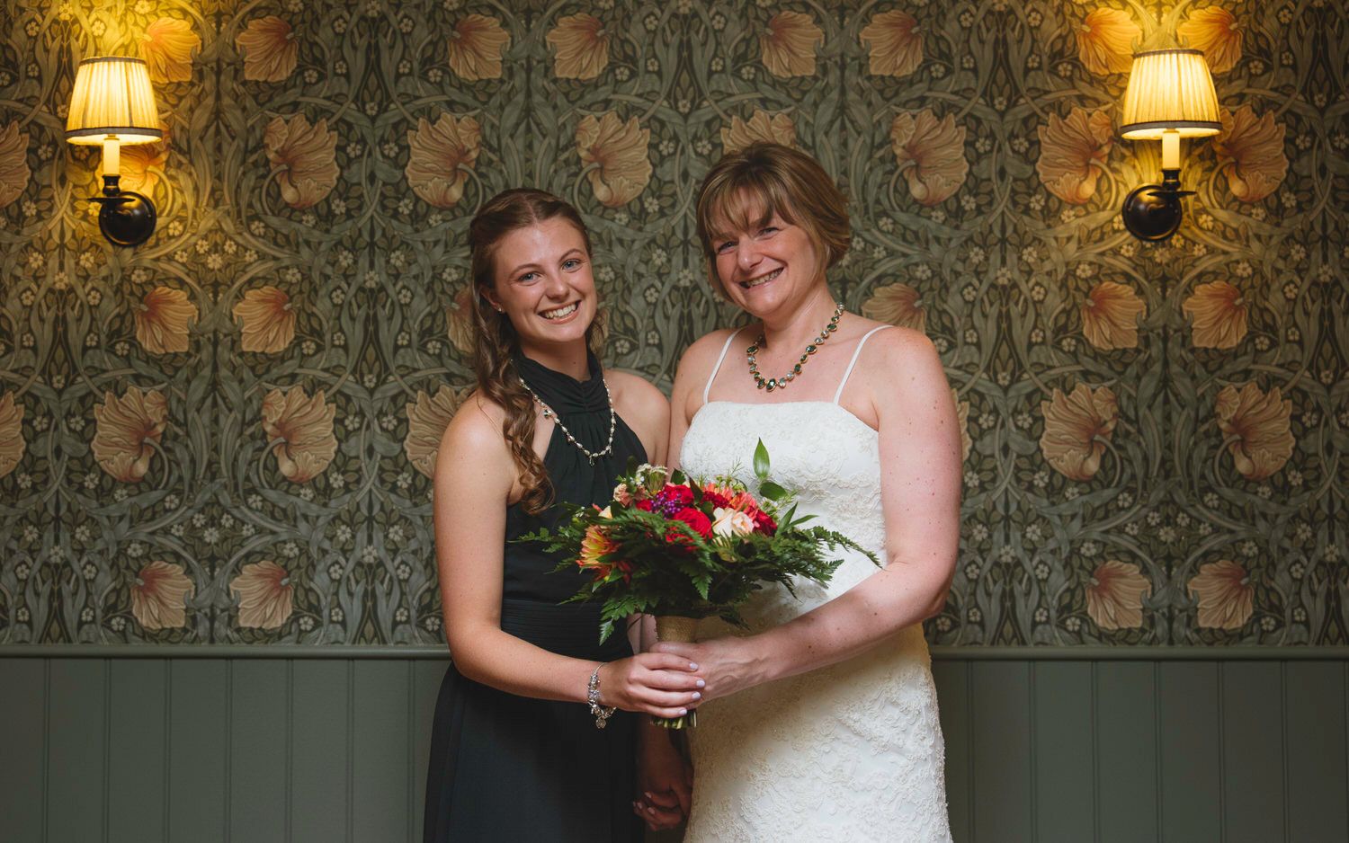 Two women pose together, one in a white wedding dress holding a bouquet, against a decorative wallpapered wall with warm lighting.