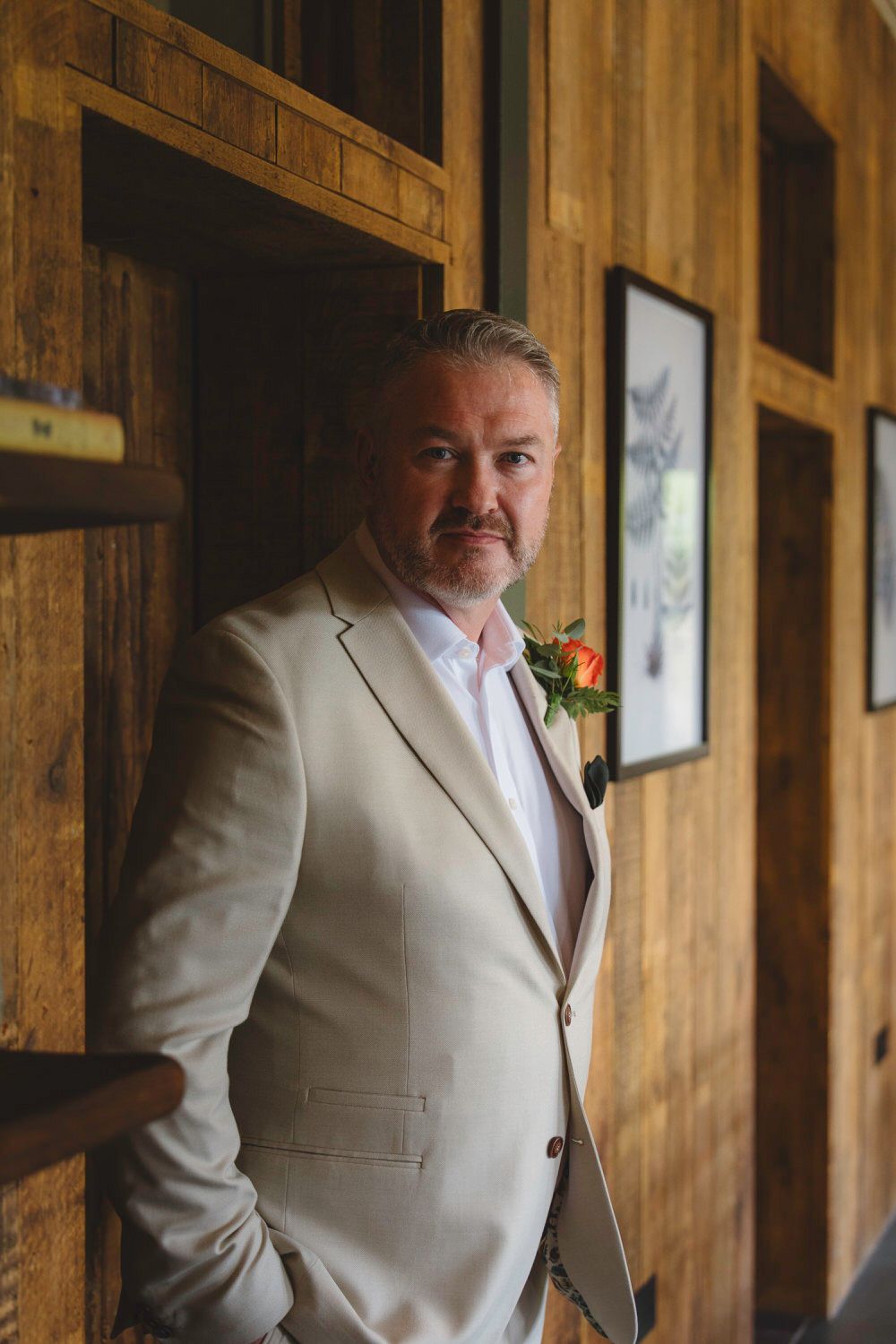 A groom in a beige suit with an orange boutonniere leans against a wooden wall, posing for a wedding portrait.