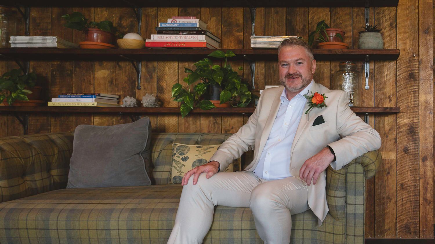 Man in beige suit with floral boutonniere sitting on plaid sofa near wooden shelves with books and plants.