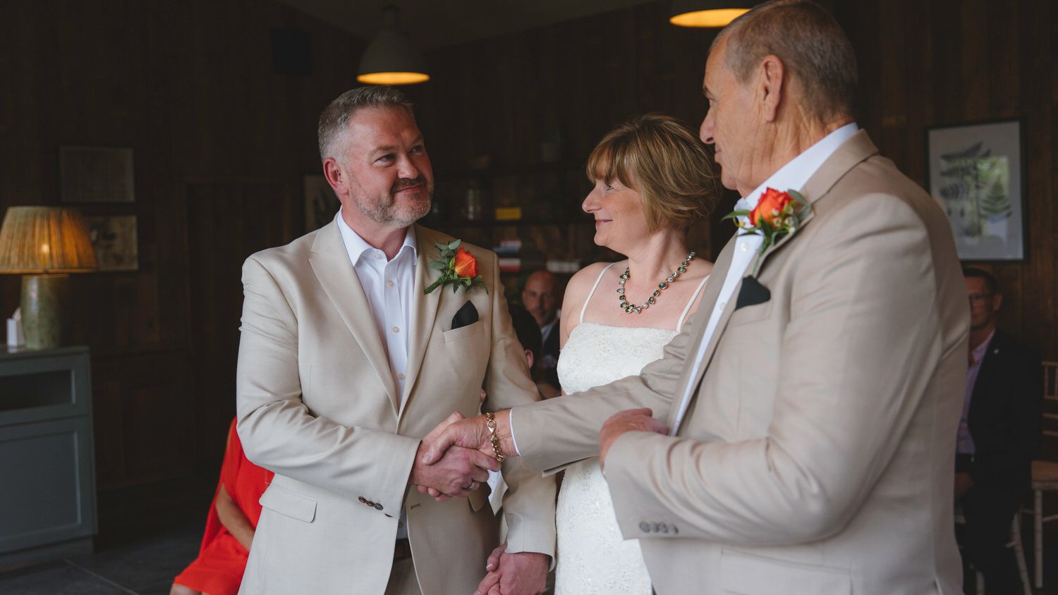Two grooms in beige suits exchange vows with a smiling bride in a white dress at an elegant indoor wedding ceremony.