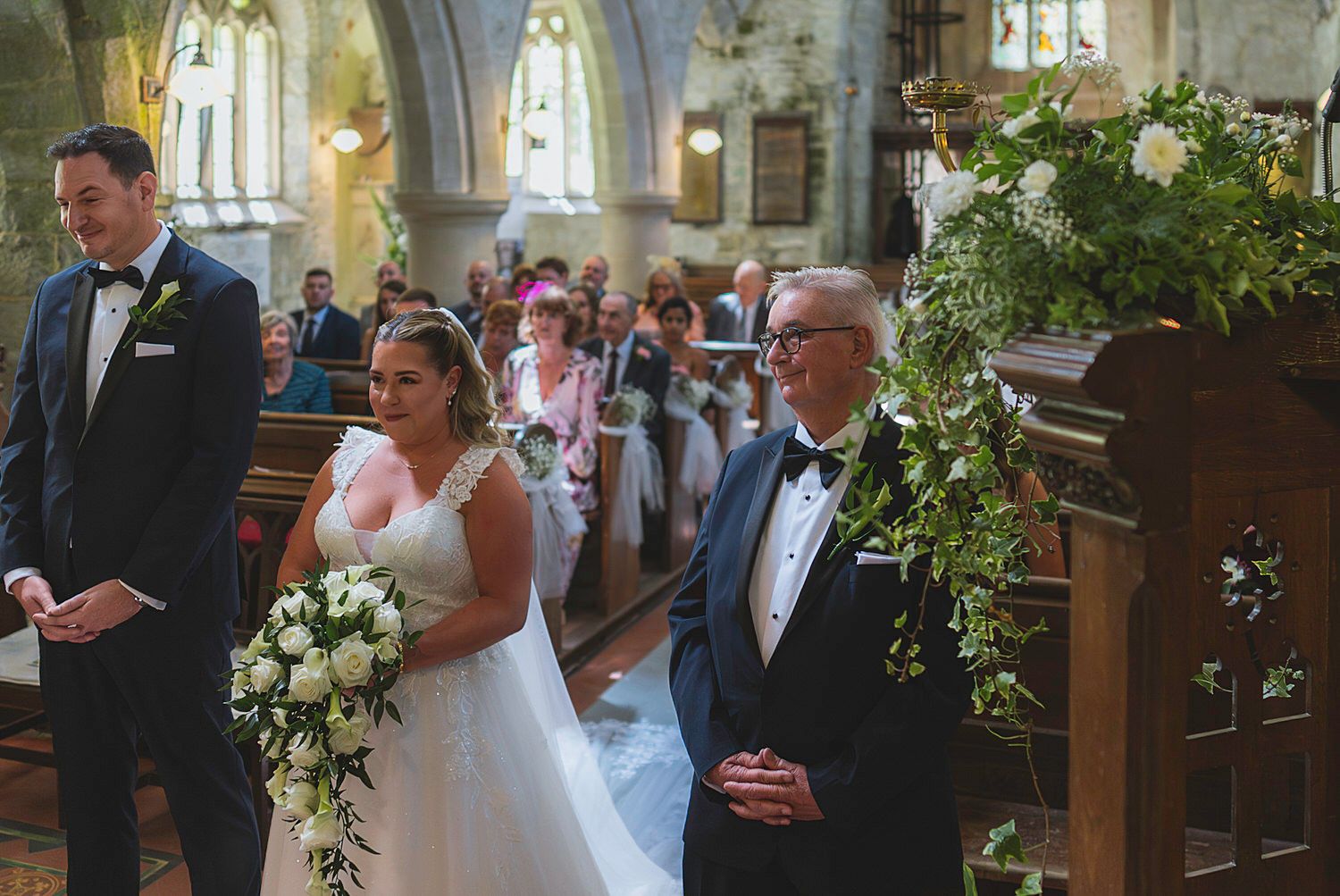 Bride walks down church aisle with escort, holding white bouquet, as groom waits at altar during wedding ceremony.