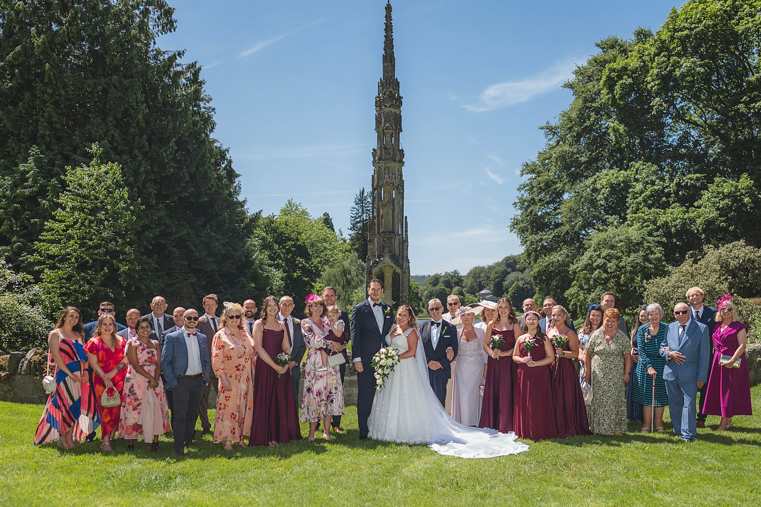 Wedding group photo with bride and groom surrounded by guests on sunny day, tall stone monument and green trees in background.