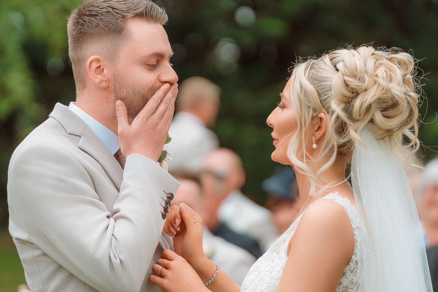 Emotional groom covers his mouth in surprise as bride smiles during outdoor wedding ceremony.