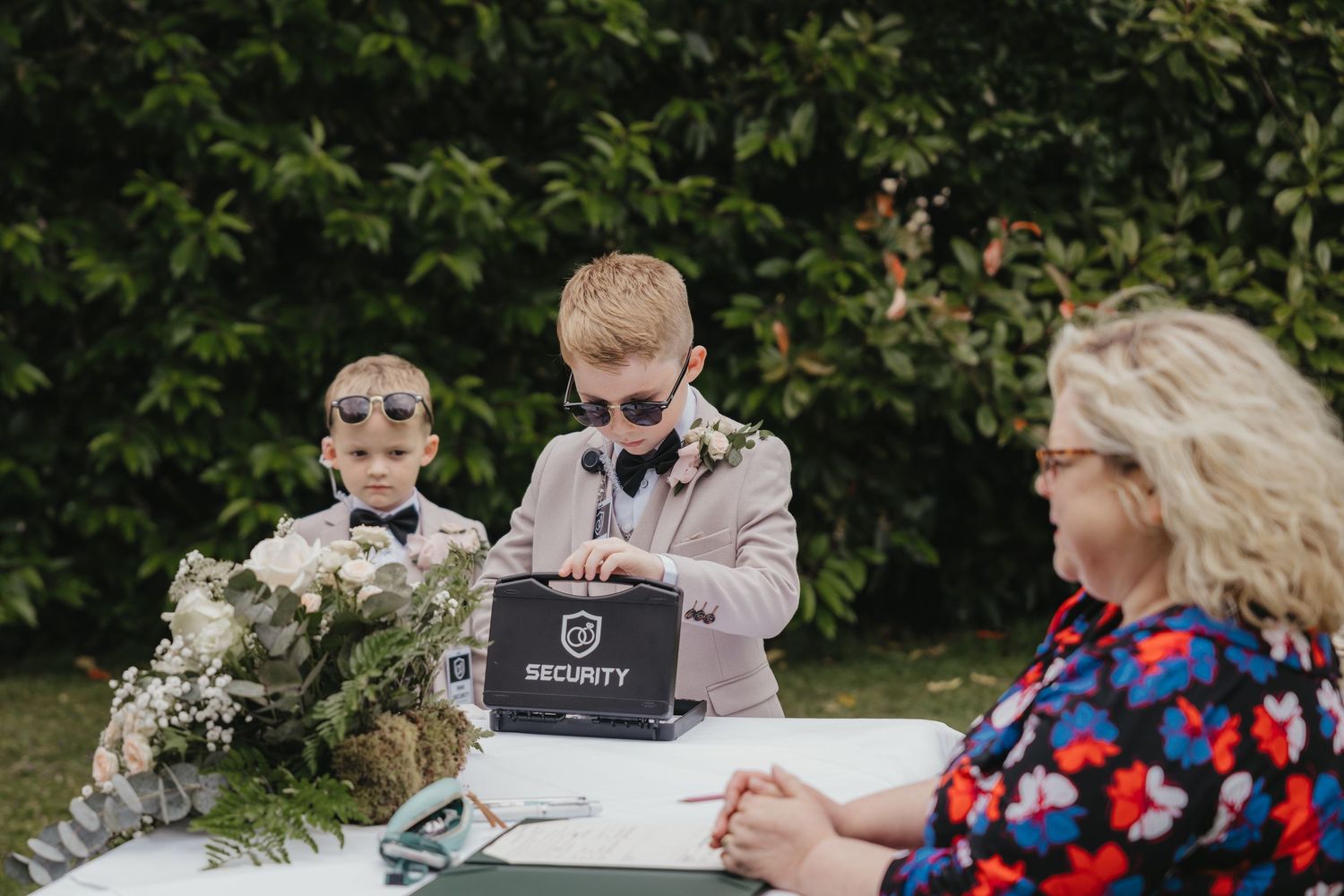 Two young boys in suits and sunglasses playing security guards at an outdoor wedding ceremony table.