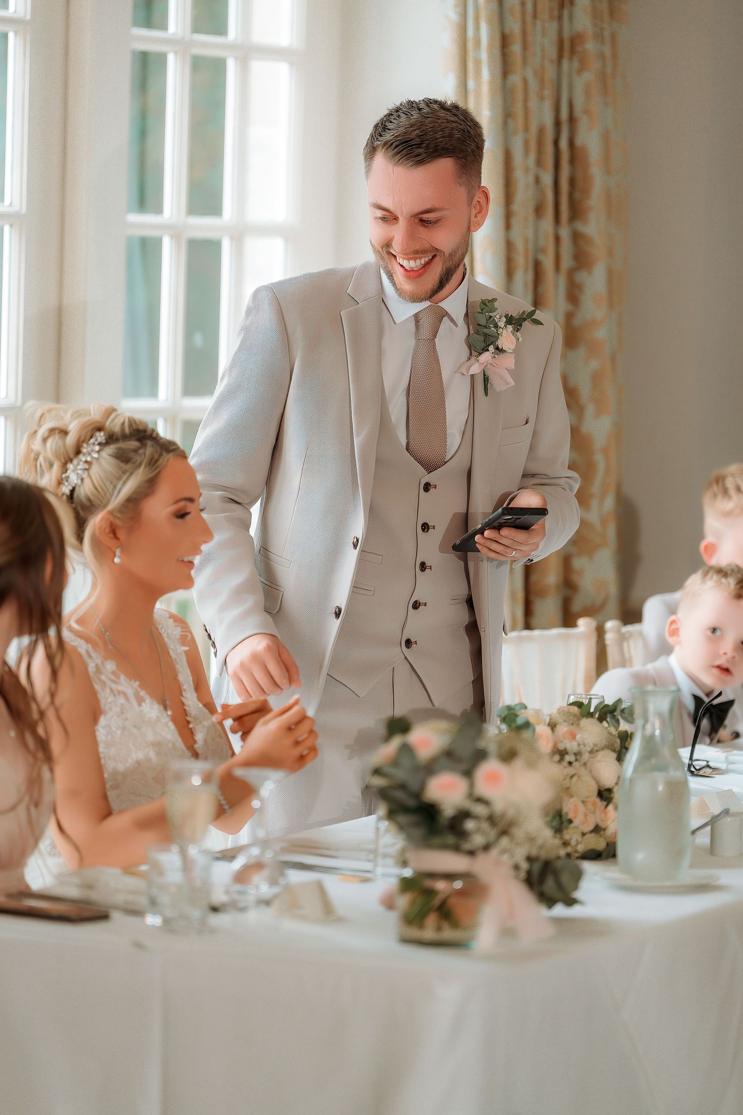 Smiling groom in gray suit holds phone at wedding reception table beside bride in white lace dress with floral centerpiece.