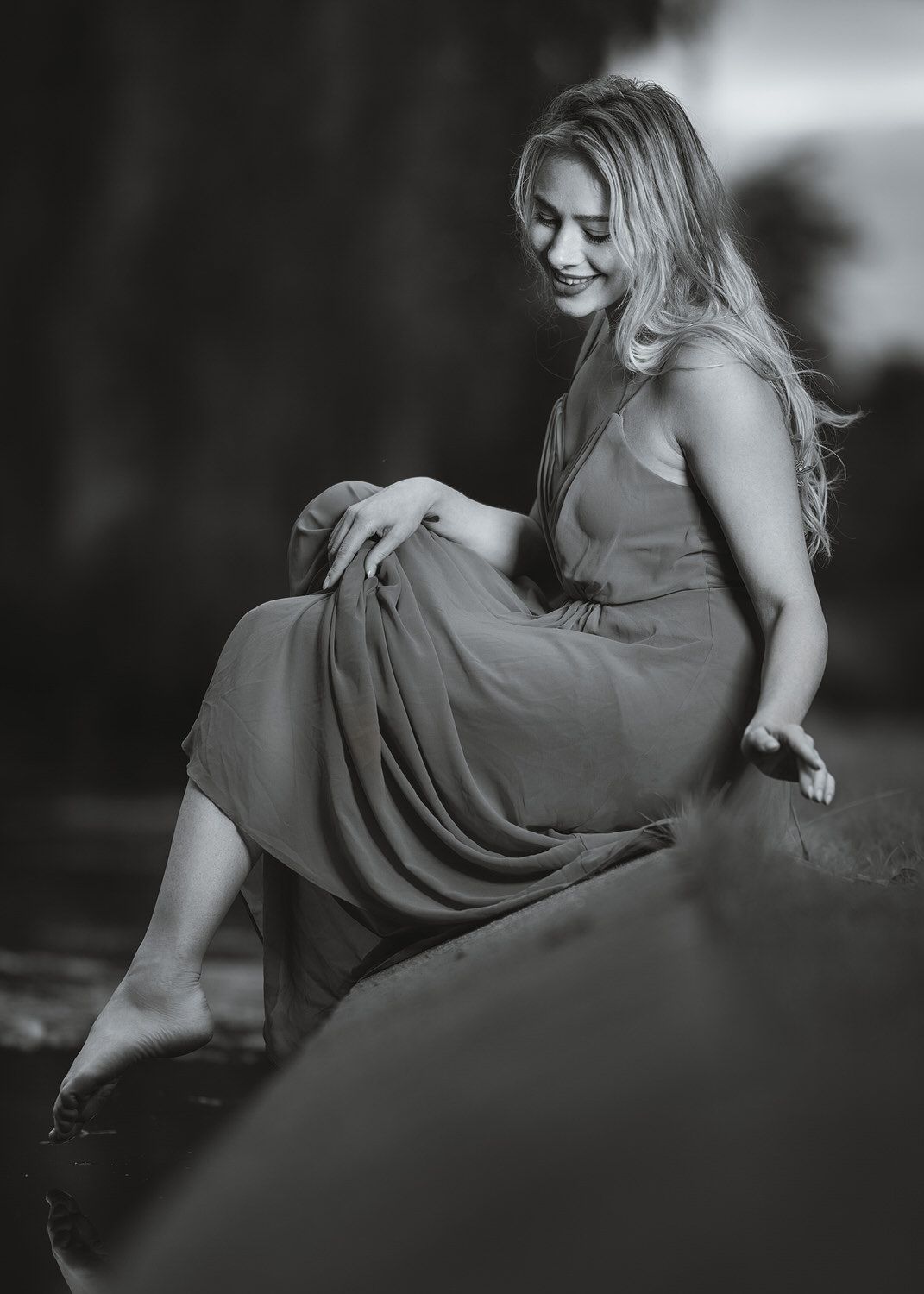 Black and white photo of a smiling blonde woman in a flowing dress sitting gracefully outdoors.
