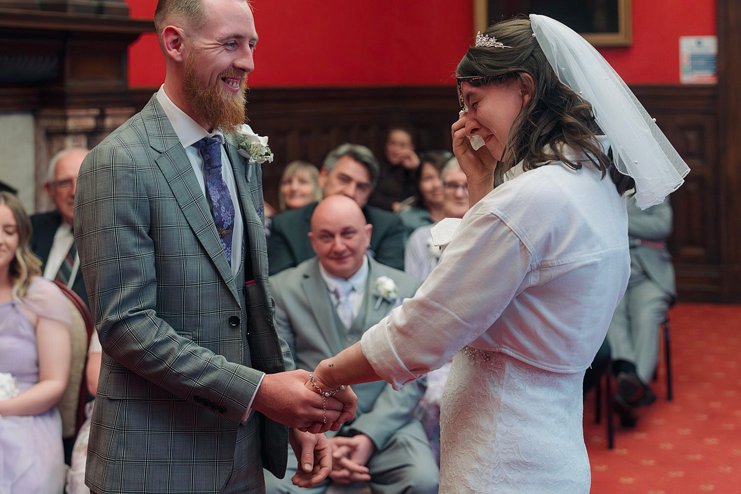 Bride and groom laughing joyfully during wedding ring exchange ceremony indoors with guests seated in background.