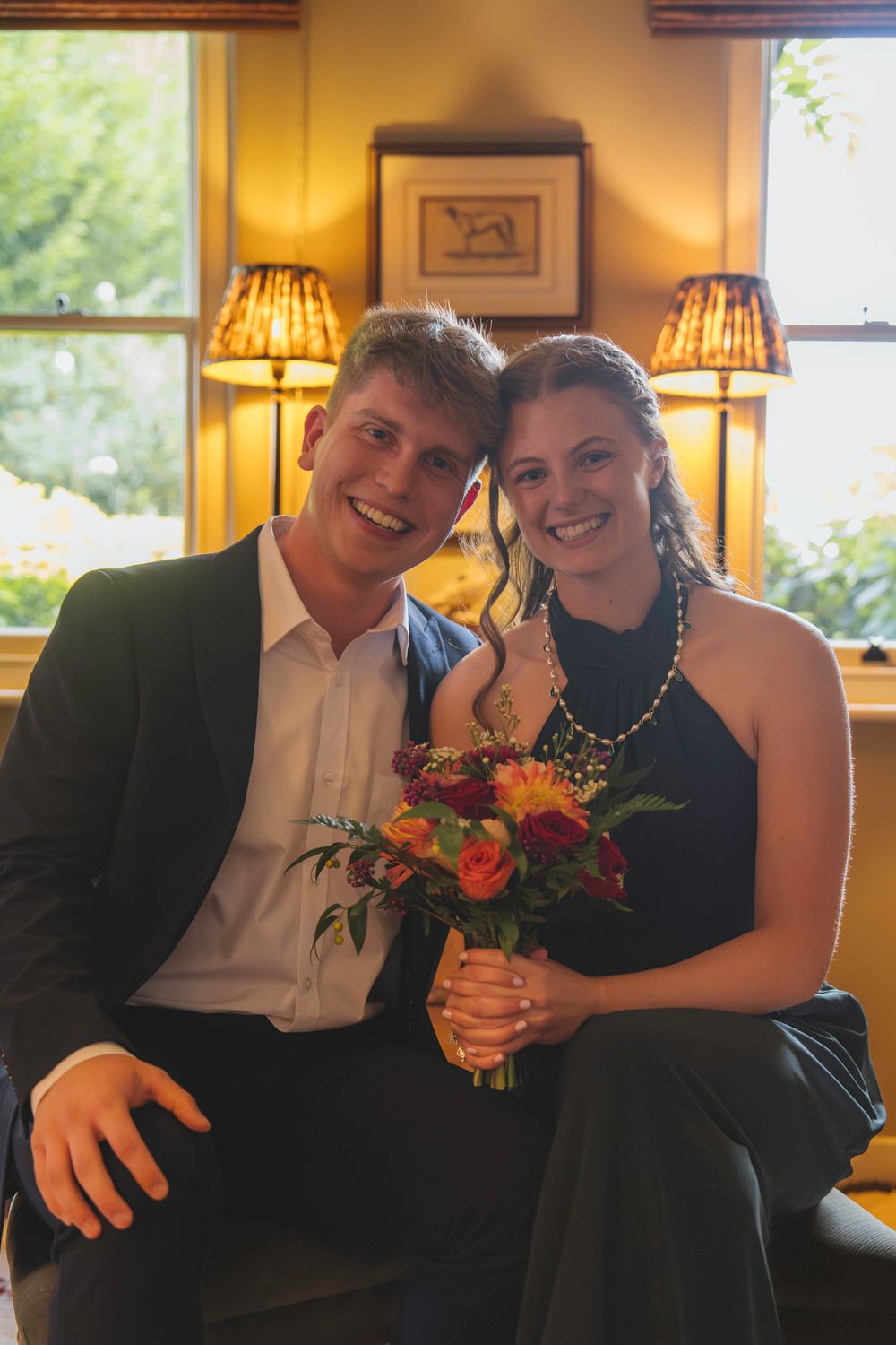 Smiling couple in formal attire holding a colorful bouquet, seated indoors with warm lamp lighting.