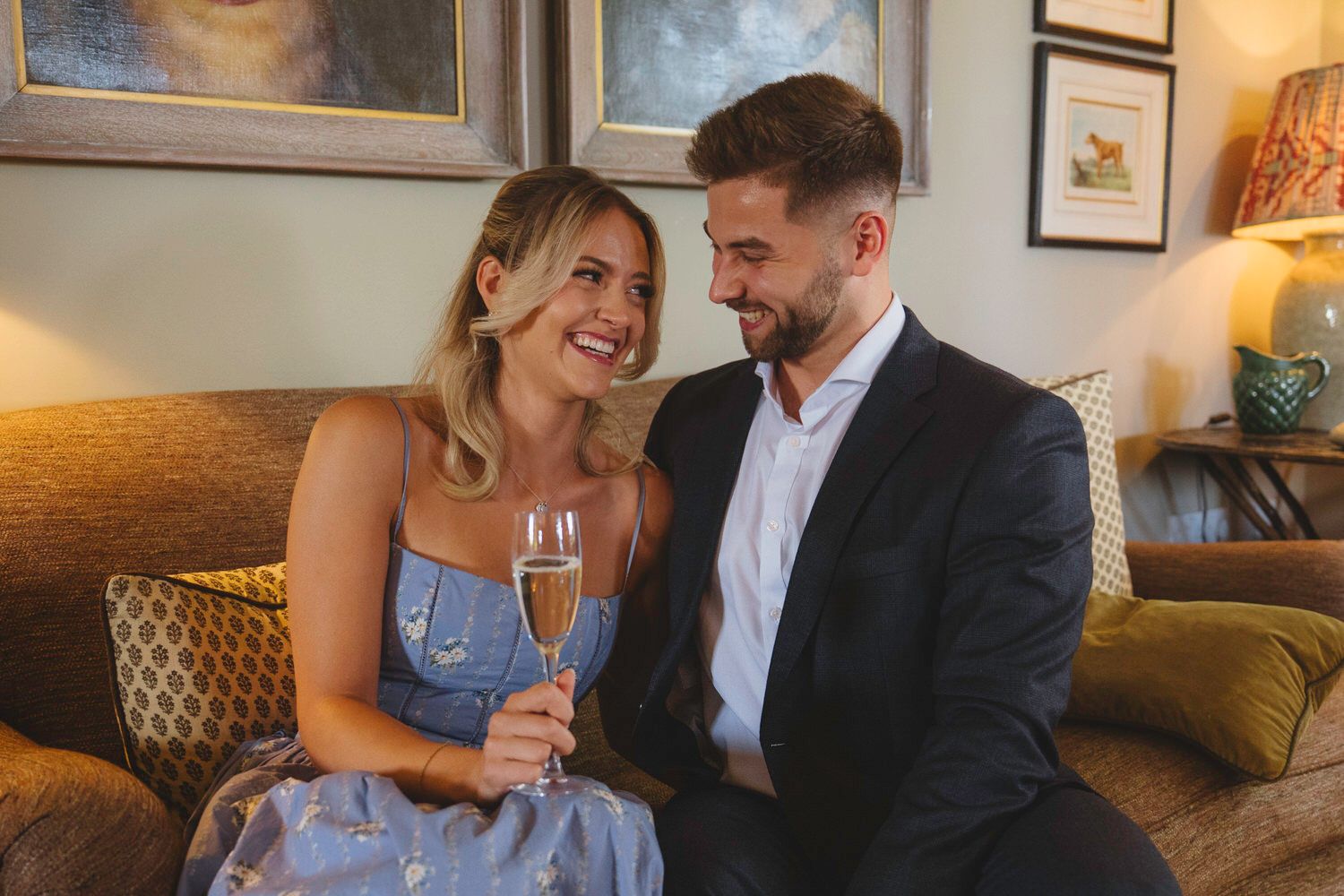 Couple celebrating with champagne on a couch, smiling at each other in elegant attire at a romantic gathering.