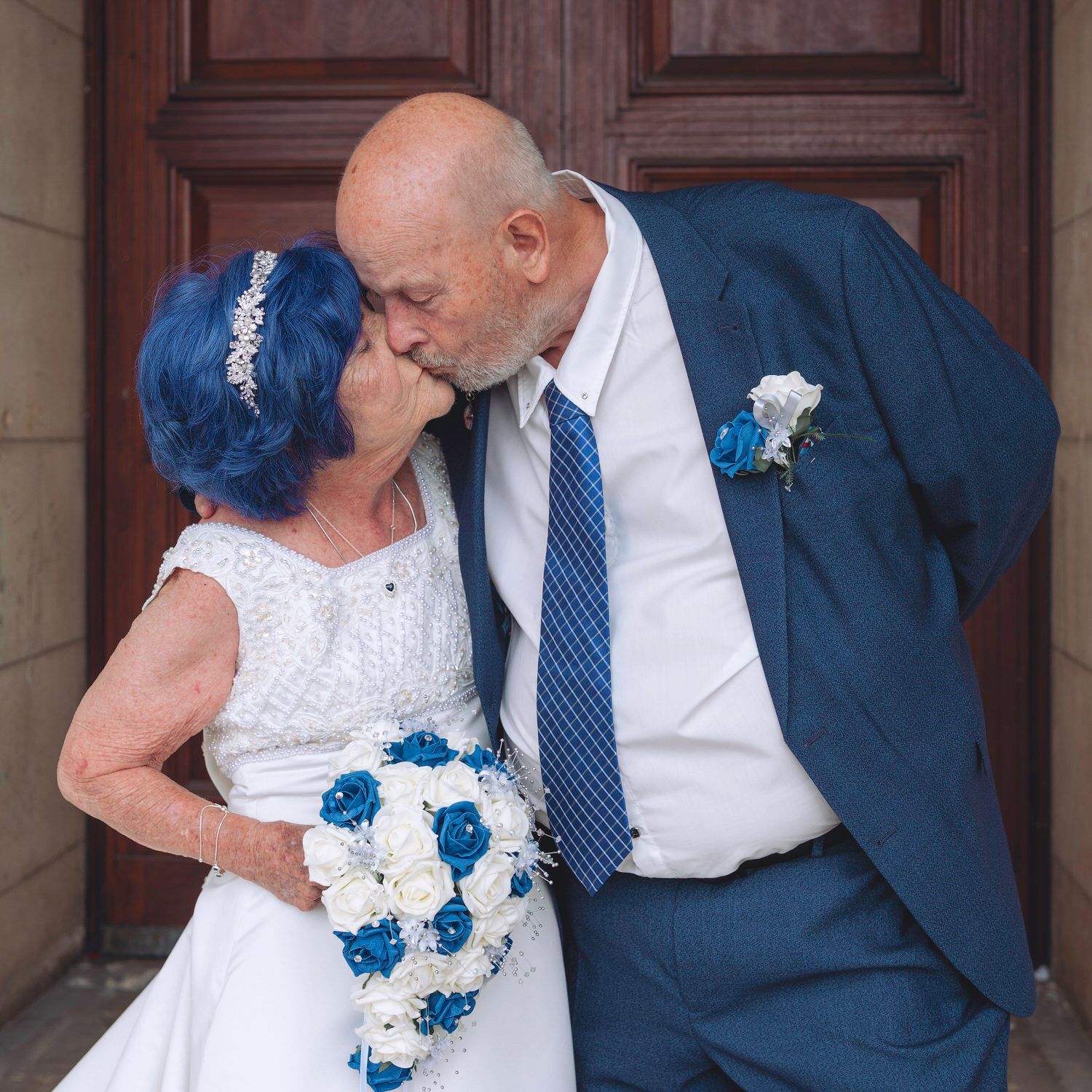 Elderly couple sharing a kiss on their wedding day, bride in white dress with blue hair holding bouquet.