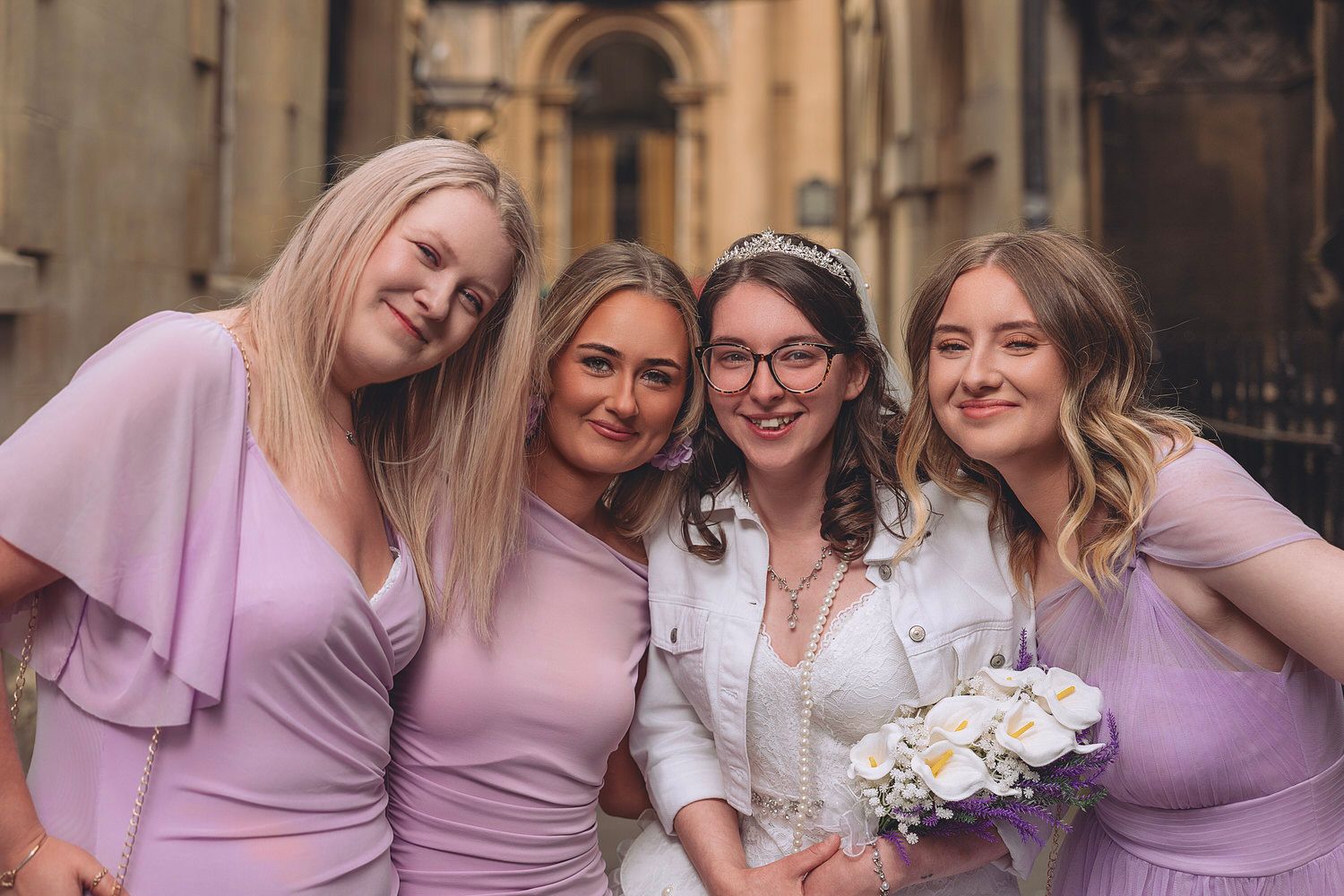 Bride in white holding bouquet poses with three bridesmaids in lilac dresses outside a stone building.