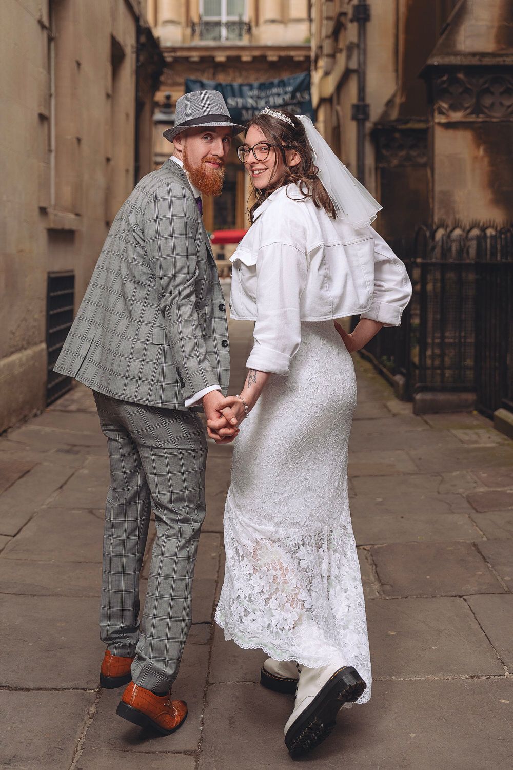 Bride and groom holding hands walking down a cobblestone alley, she in white lace gown, he in gray suit.