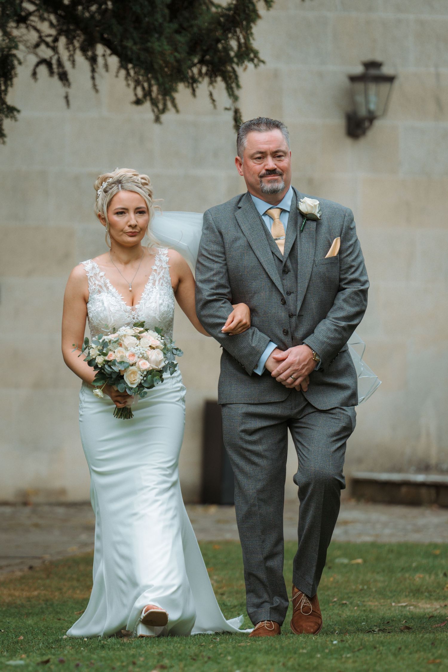 Bride in white gown holding bouquet walks with suited man at outdoor wedding ceremony.