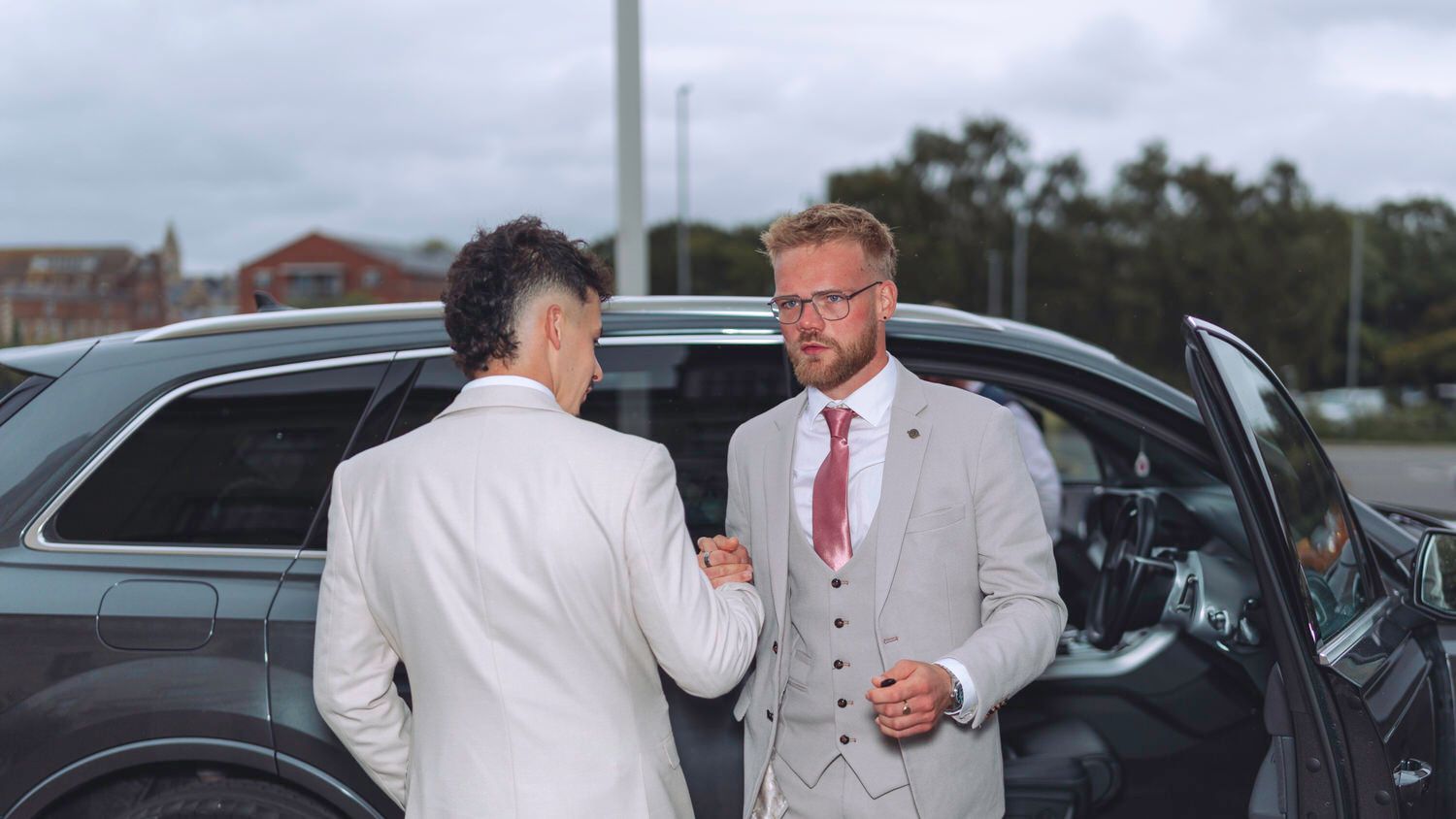 Two men in suits shaking hands beside a dark car outdoors.