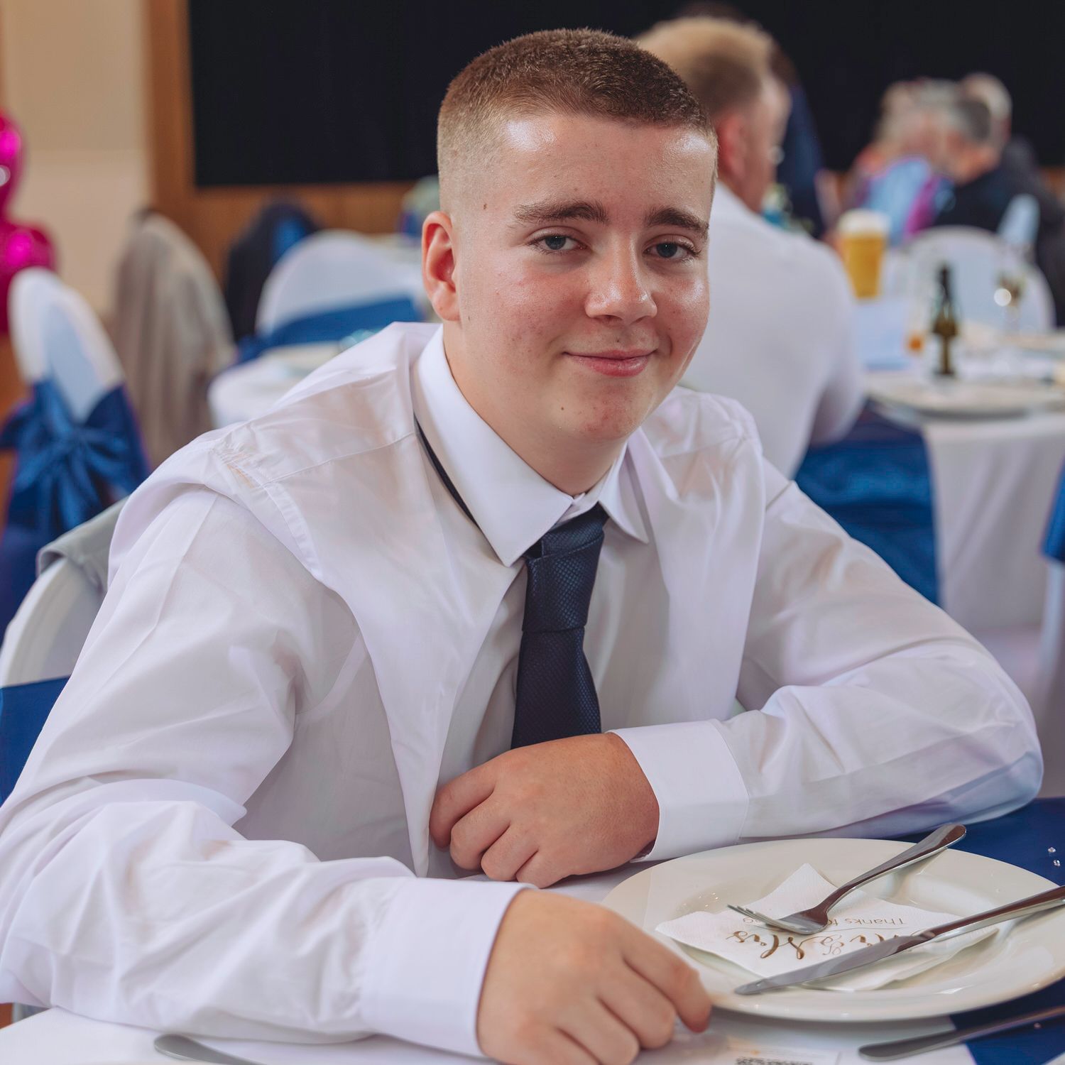Young man in white shirt and navy tie smiling at a formal banquet table with blue chair covers.