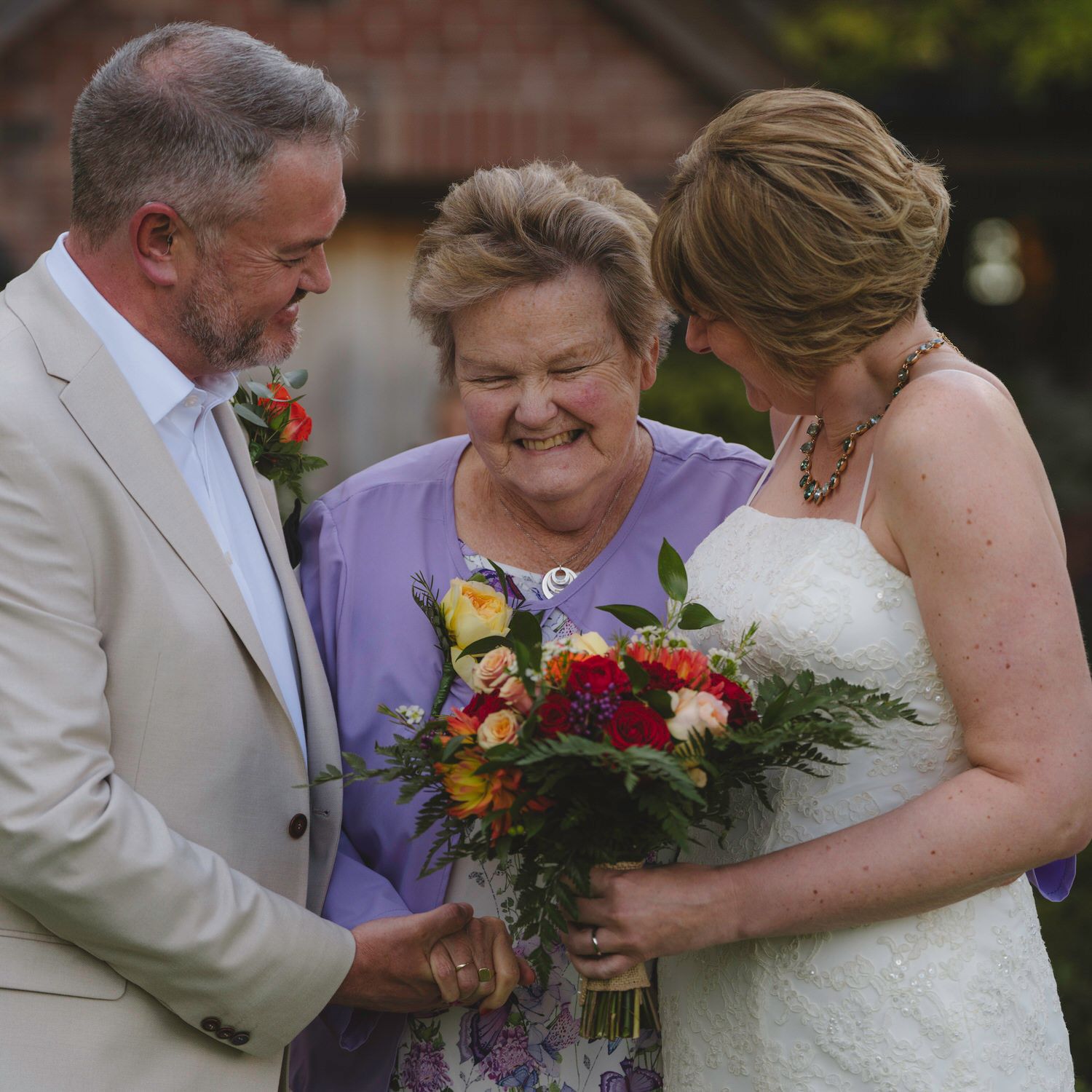 Bride and groom share a joyful moment with an older woman holding a colorful bouquet at an outdoor wedding.